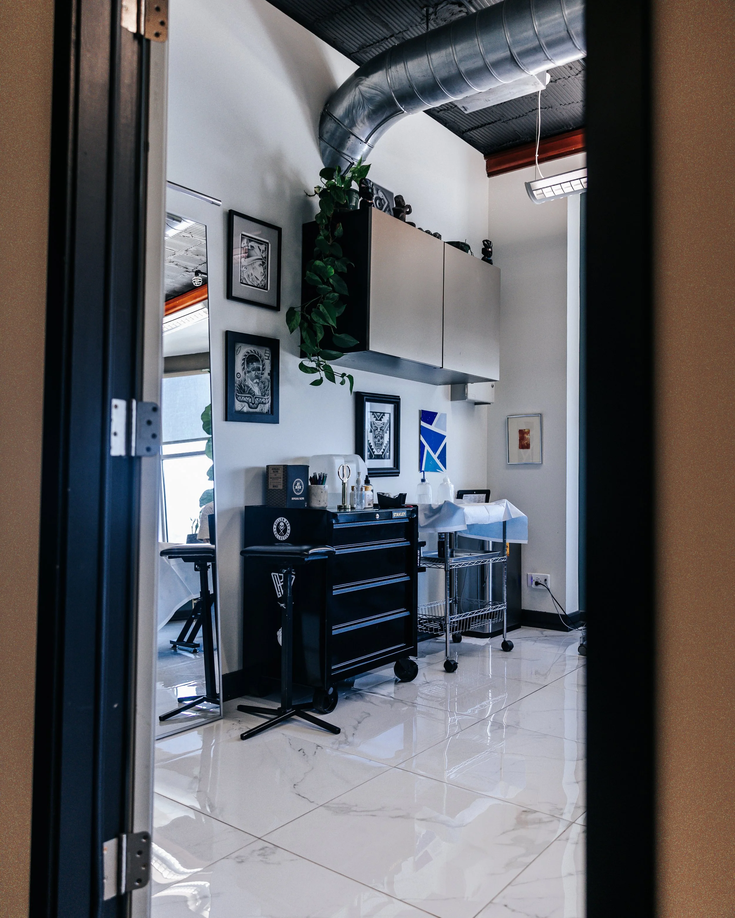 View of a modern indoor space through a partially open door. The room features a white wall with framed art, a black cabinet with various items, a rolling cart, and artistic decor on the wall. There's ductwork running along the ceiling, and the floor