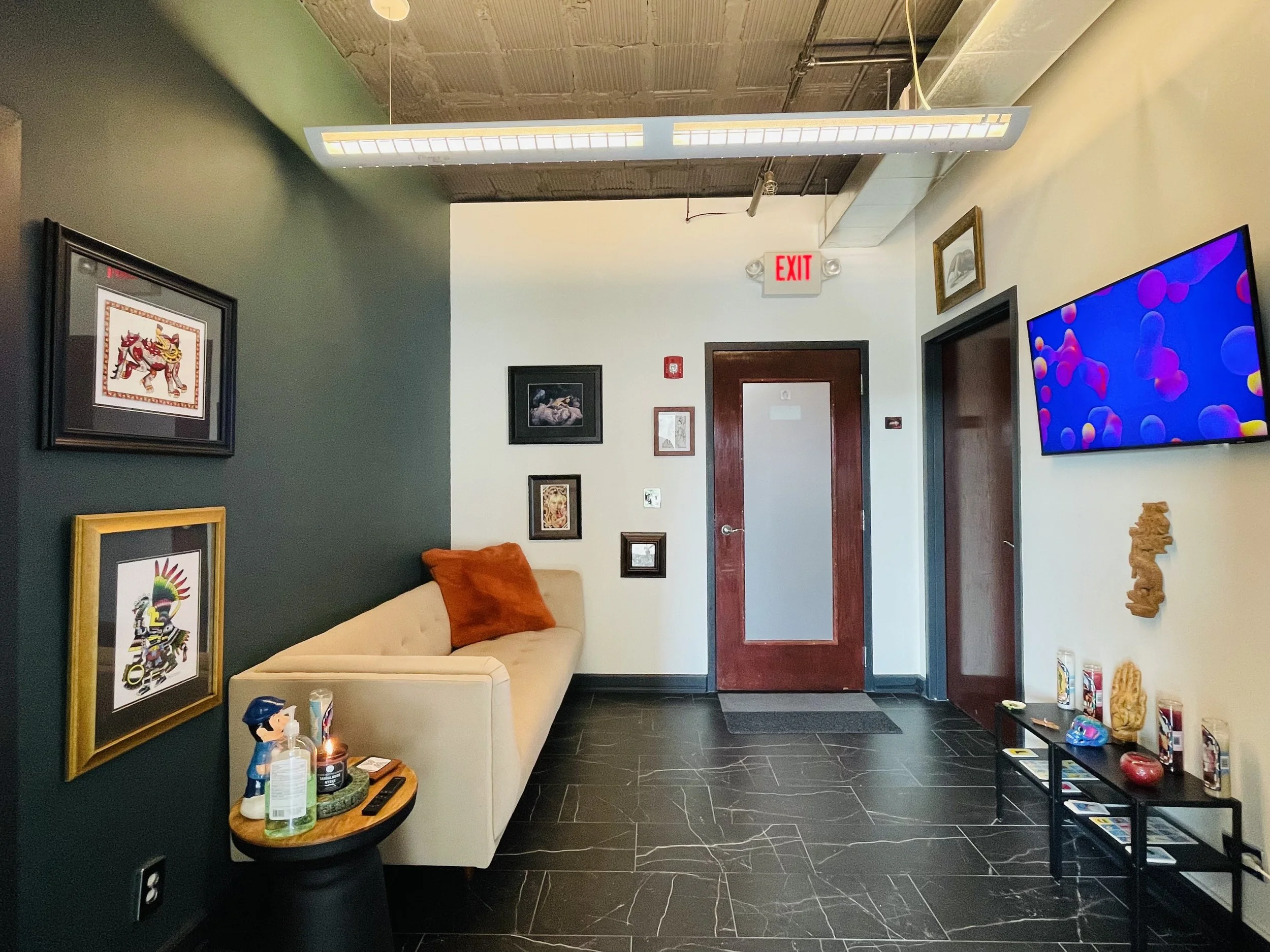 Interior of an art gallery or waiting area with a beige sofa, orange pillow, wall-mounted TV, artwork on walls, and decorative objects on a small black table and shelves.