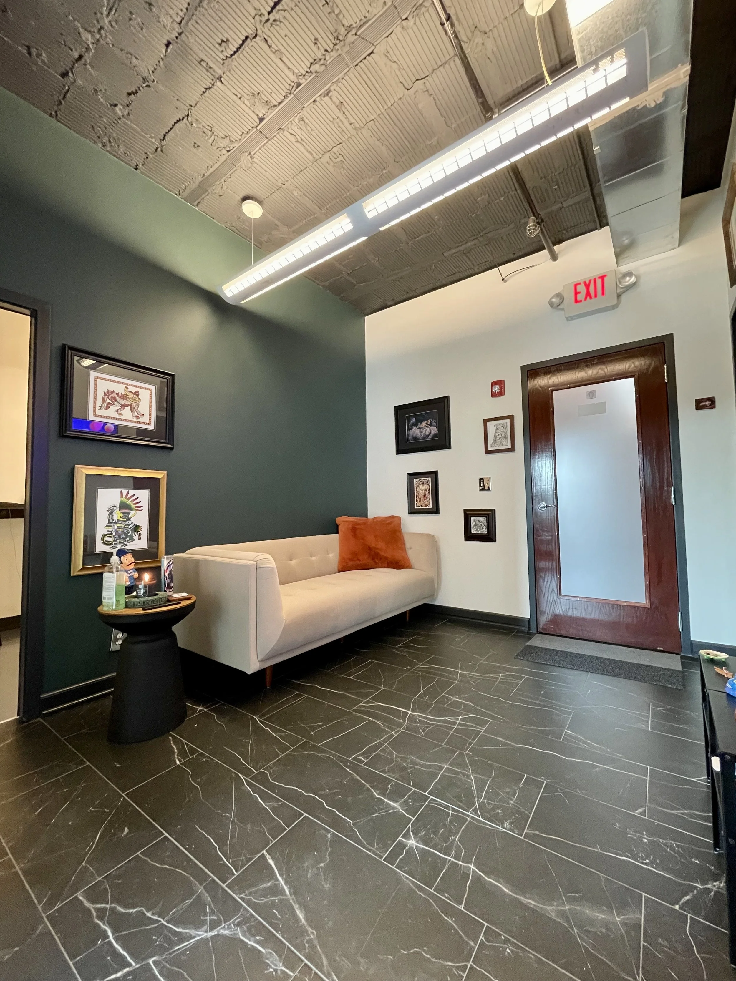 A modern indoor waiting area with black tiled floors, a cream couch with an orange pillow, black framed art on the walls, a black side table with hand sanitizer and a candle, and a wooden door with a glass panel, illuminated by a false ceiling with e