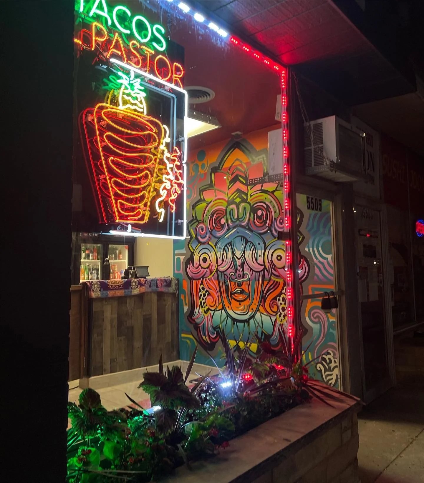 A neon sign advertising tacos and pastor, with a colorful mural of an ornate, stylized face wearing a crown, in front of a taco restaurant at night.
