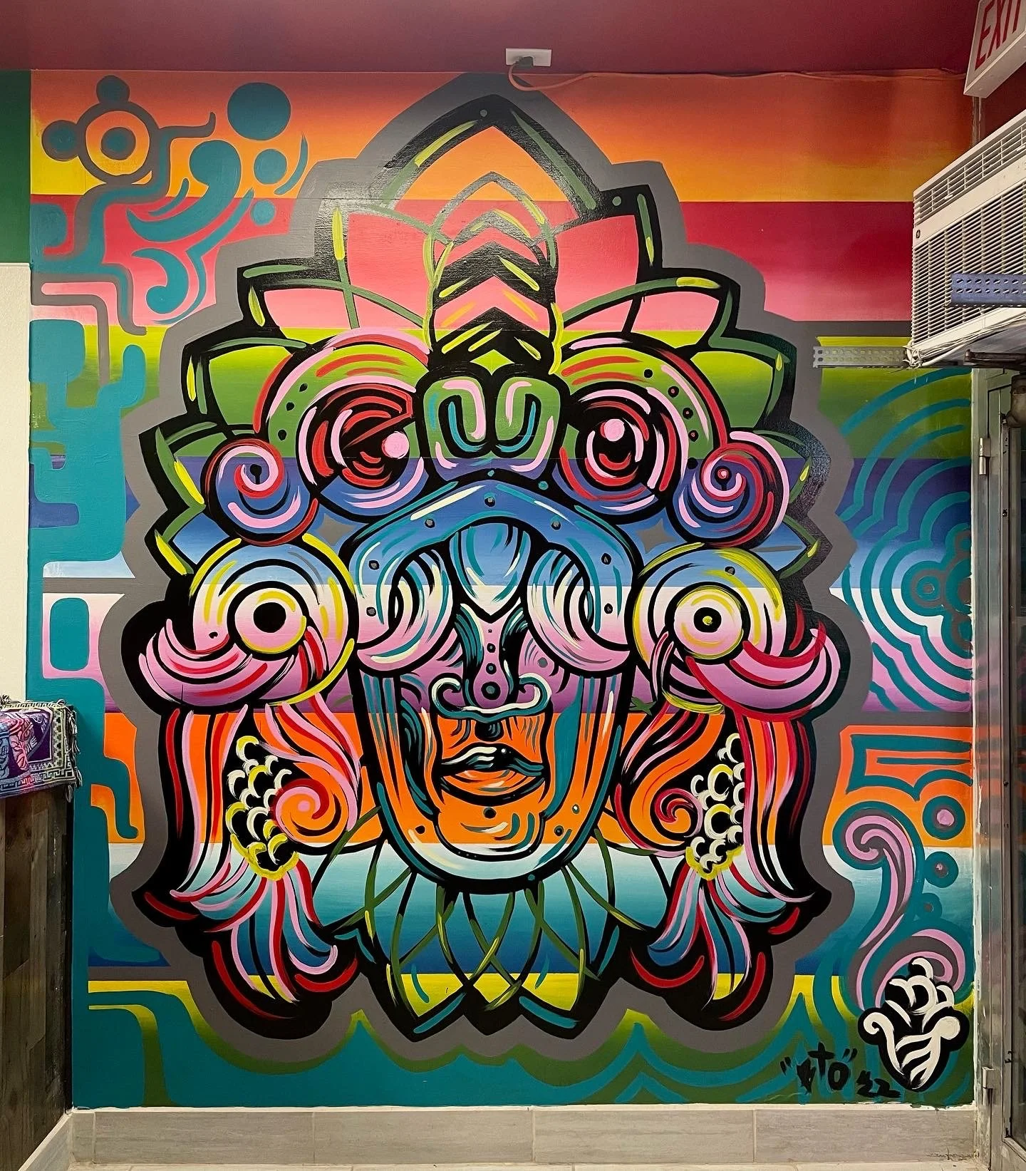 Colorful mural of a stylized face with intricate patterns, swirls, and shapes in pink, blue, green, yellow, orange, and black on a wall.