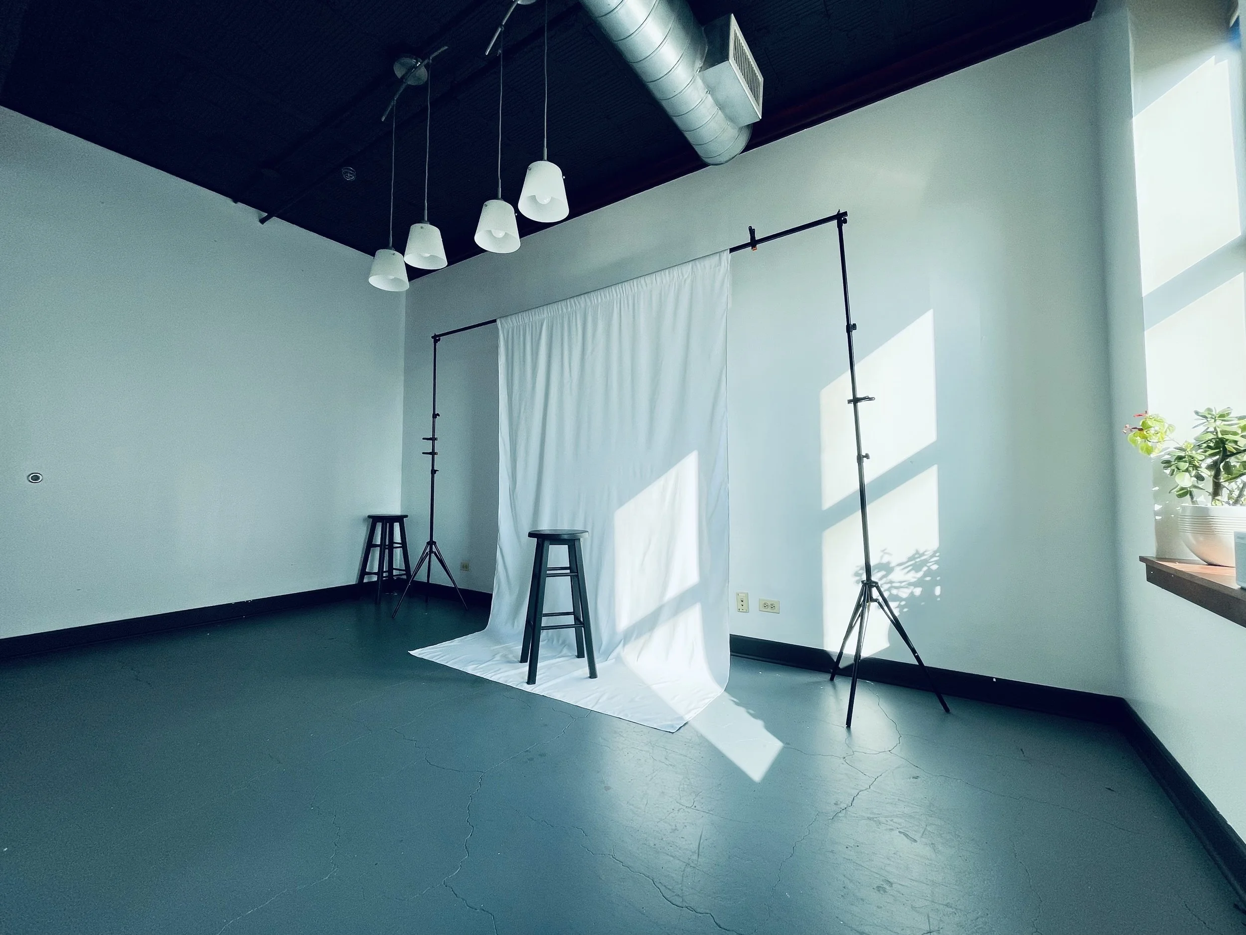 Photography studio with a white backdrop, two black stools, and lighting equipment, sunny daytime with natural light coming through the window, indoor setting