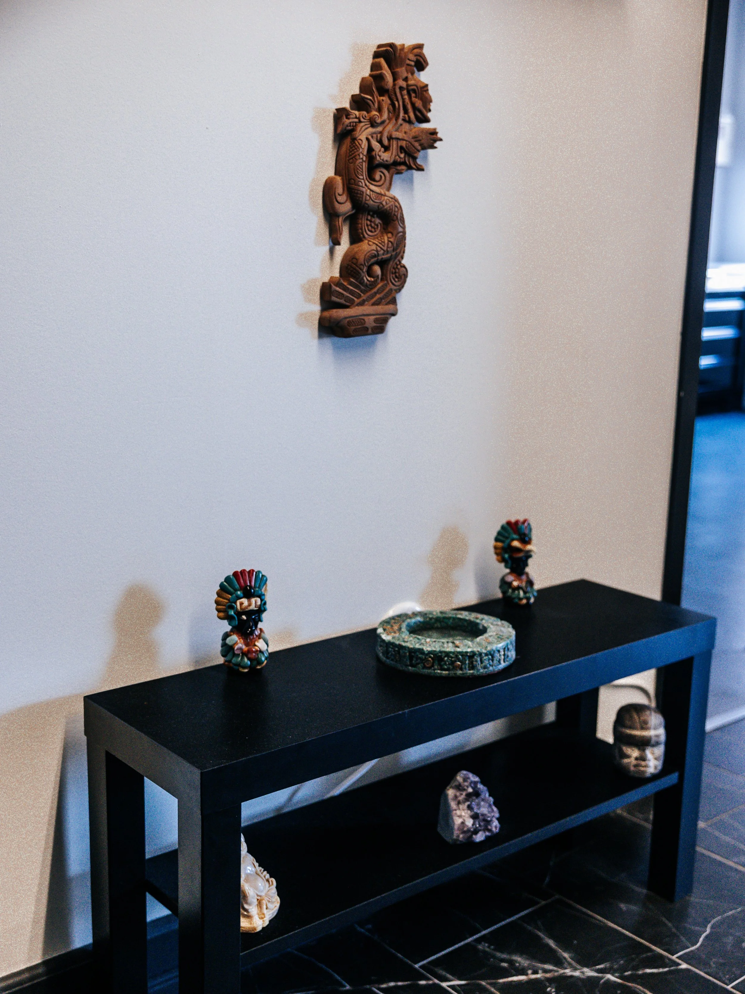 Decorative display with a carved wooden lion wall hanging, two small colorful tribal figurines, a green ring-shaped artifact, and various stones on a black table.