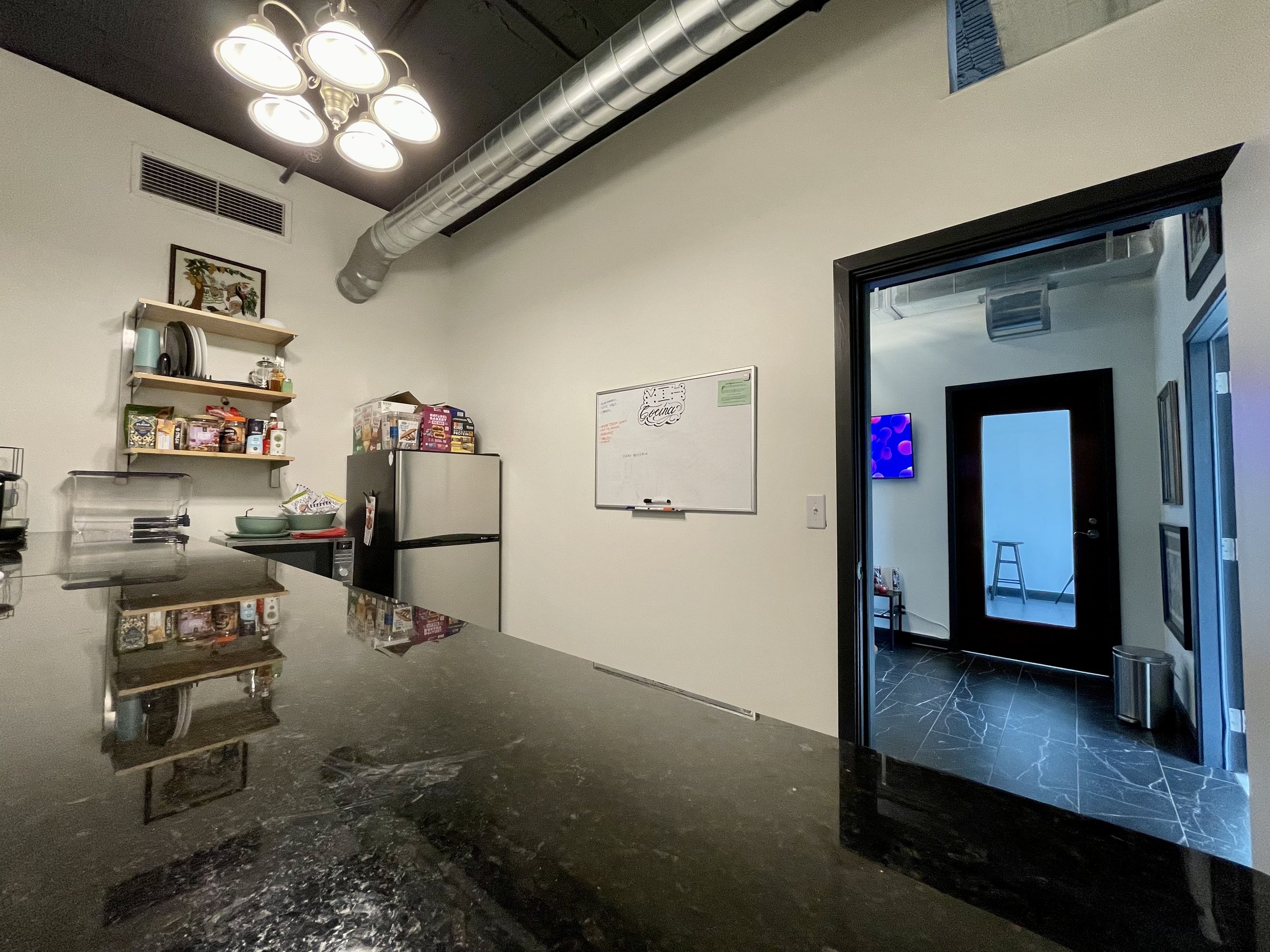 Interior of a kitchen with a black marble counter, a refrigerator, open shelves with dishes and food items, a whiteboard on the wall, and a doorway leading to a room with a black door, blue lighting, and a stool.