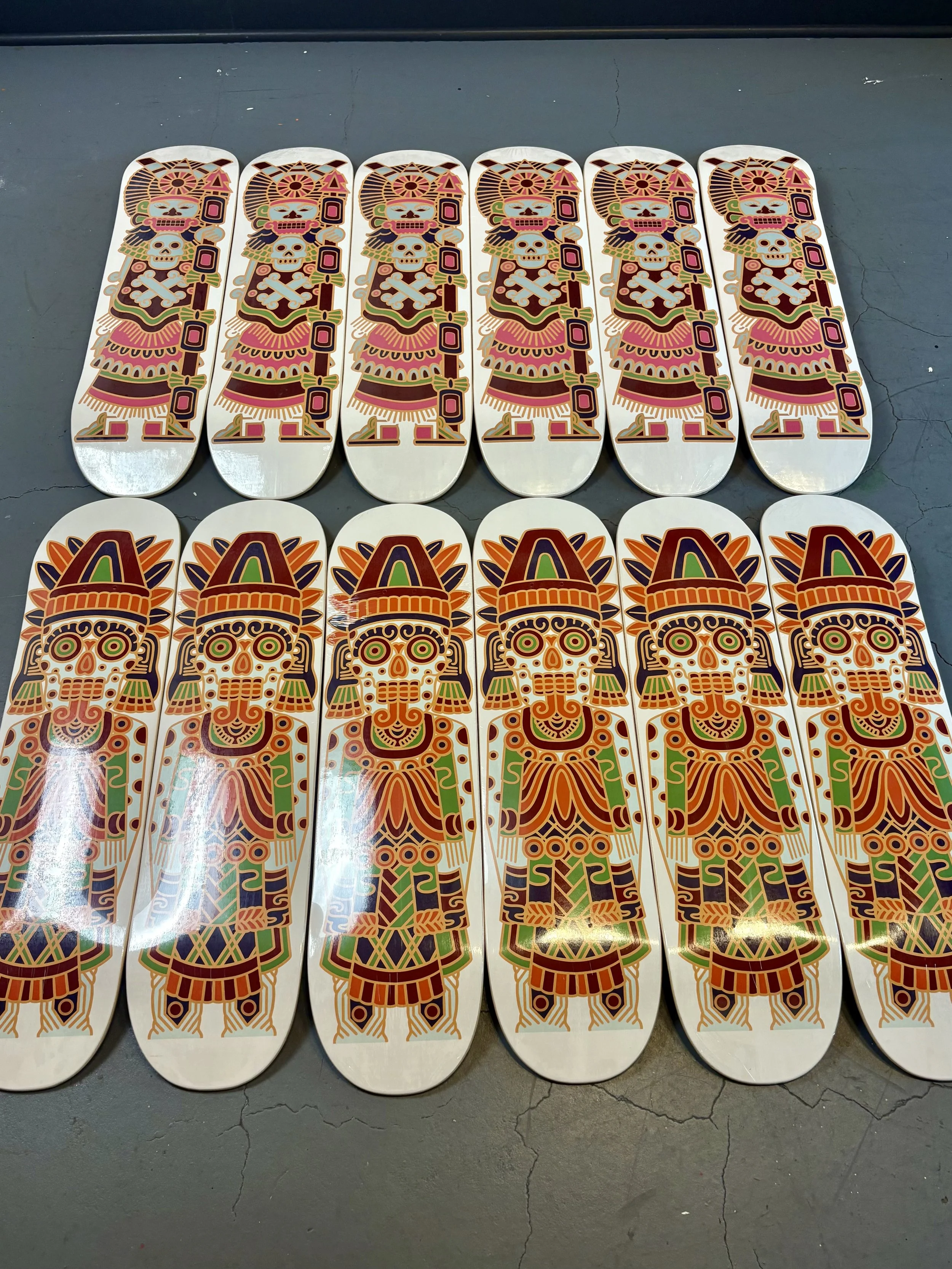 Sixteen skateboards with colorful Mayan or Aztec-style artwork arranged on a concrete floor.