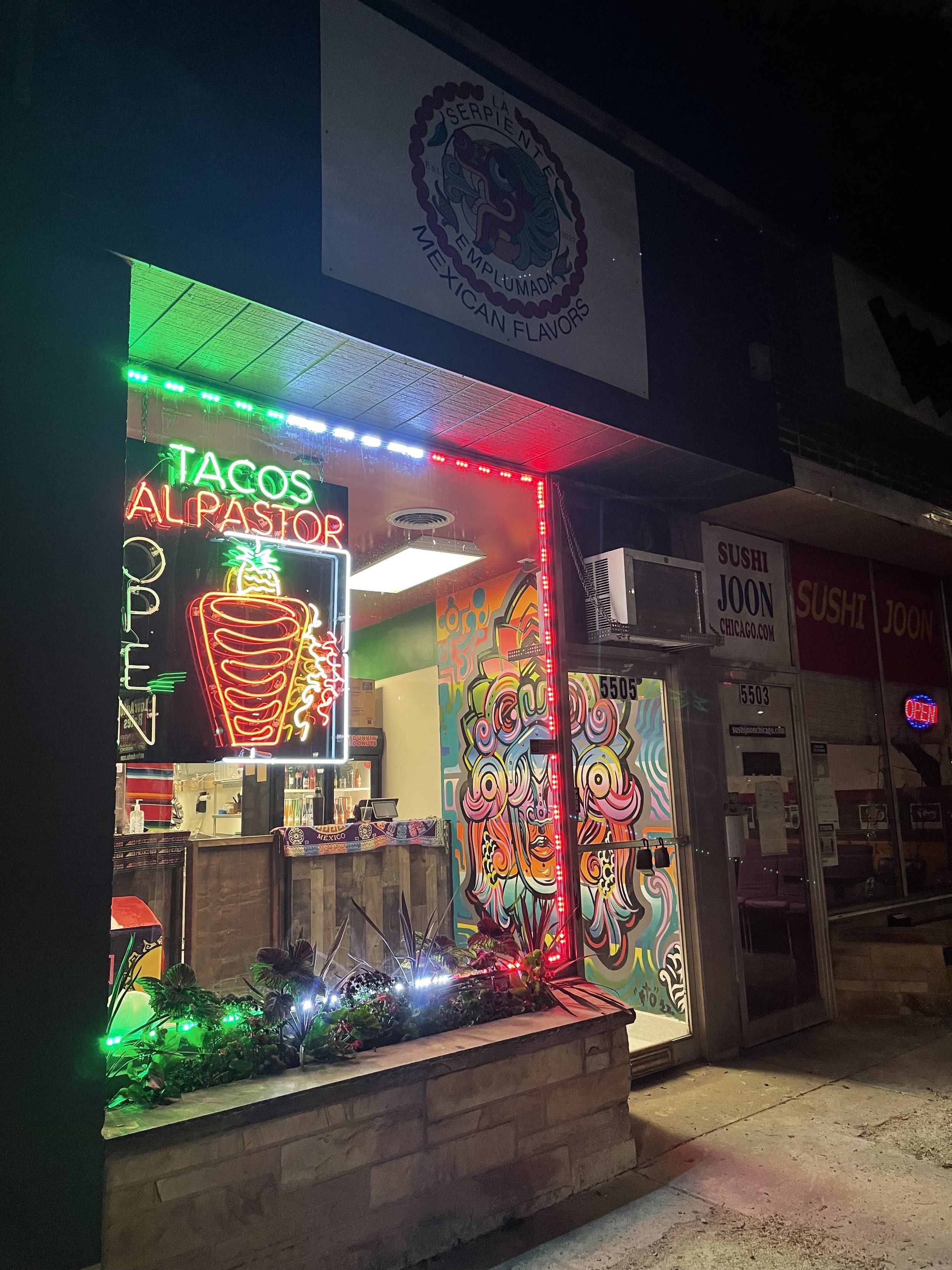 Neon sign outside a restaurant with colorful mural art inside, advertising tacos al pastor, with restaurant decor and a nighttime street scene.
