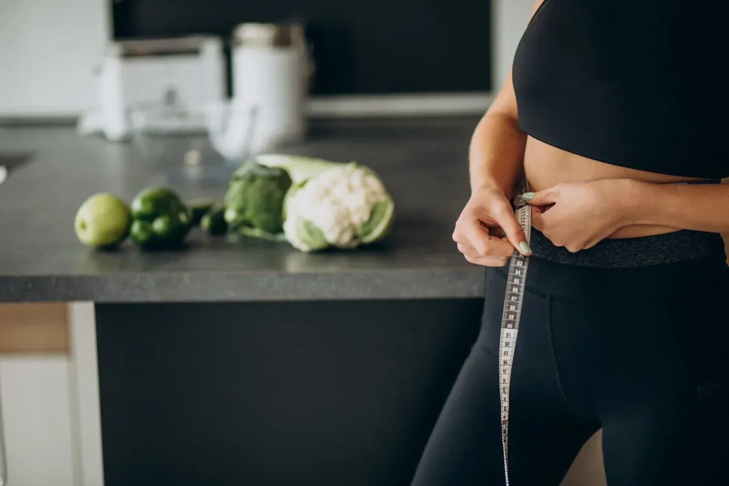A woman in athletic wear measures her waist with a tape measure in a kitchen.