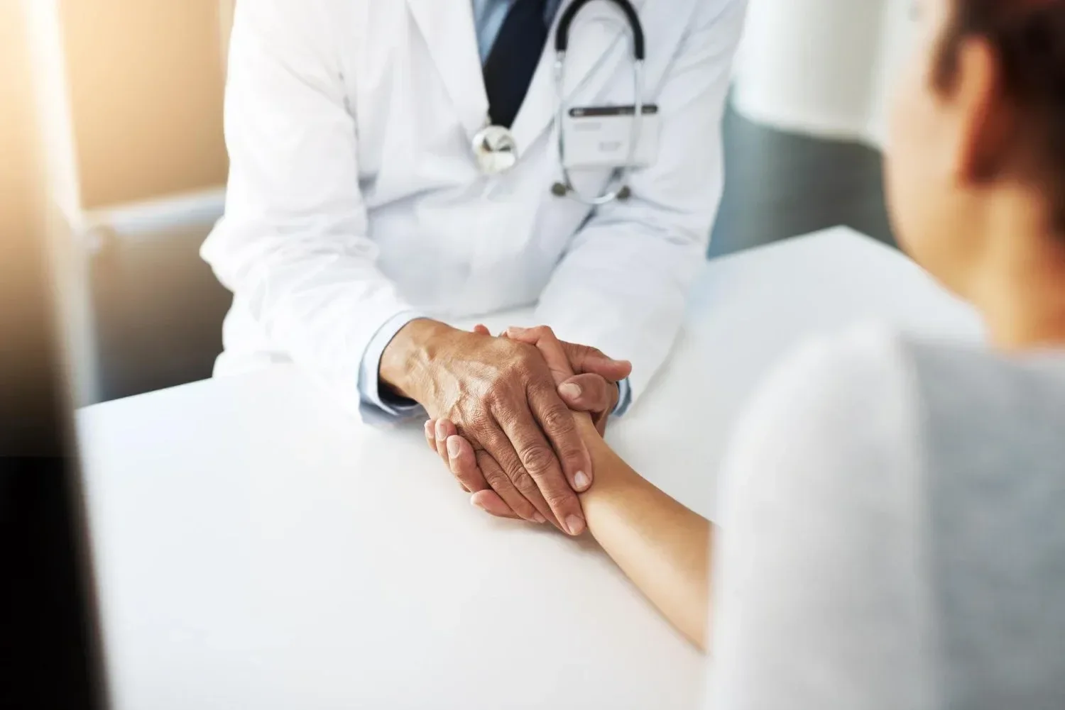 A doctor in a white coat gently holds a patient's hand across a table, conveying empathy and support.