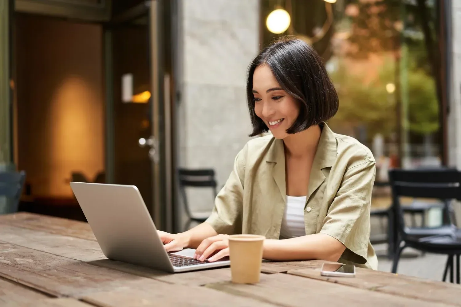 A woman with short dark hair smiles while typing on a laptop at an outdoor café.