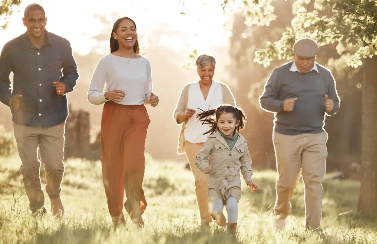 A family of five, including a child and adults, joyfully run through a sunlit park. 