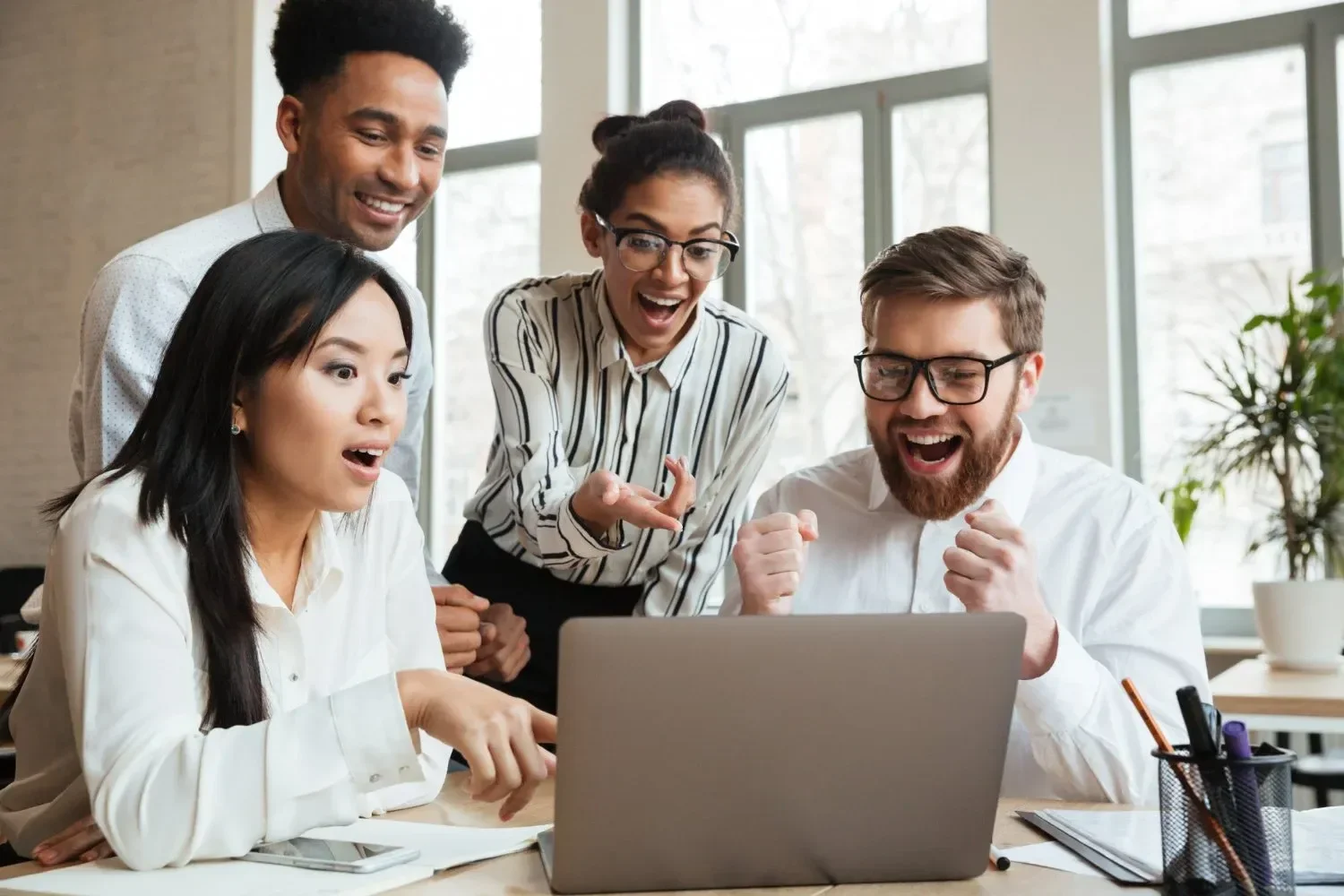A diverse group of four colleagues excitedly celebrates a success, gathered around a laptop in a bright office space.