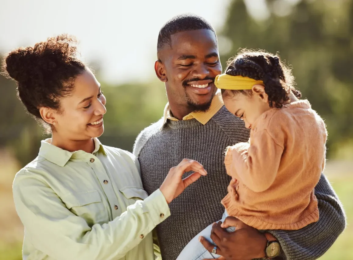 A smiling family stands outdoors. A man holds a young girl as a woman gently touches her.