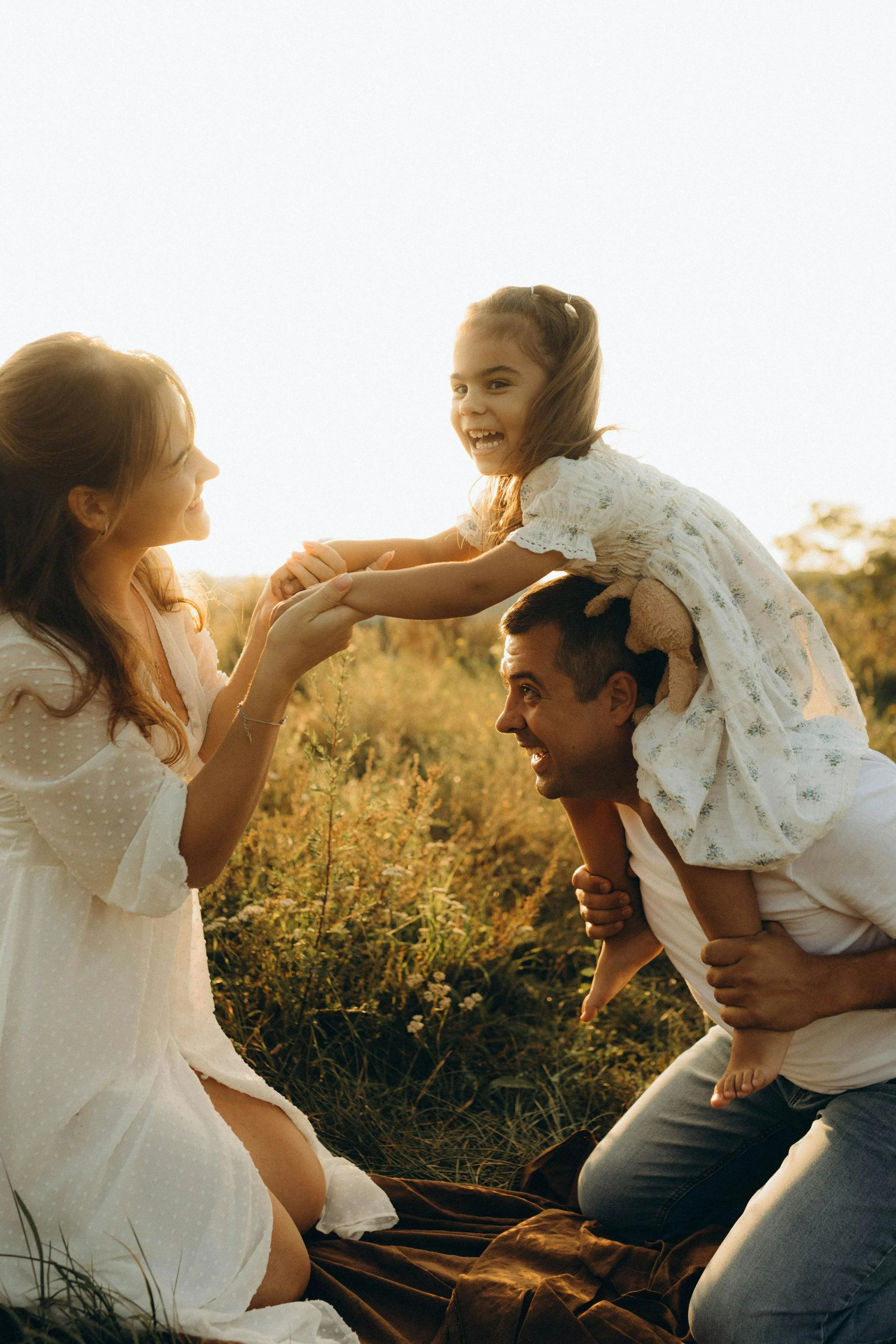 Family of four outdoors during sunset, father giving girl a piggyback ride, enjoying a playful moment.