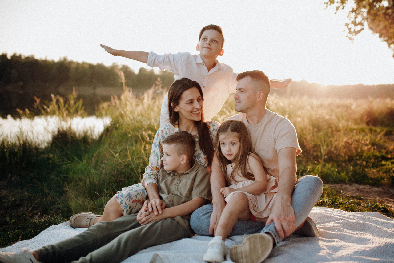 Family of six sitting on a blanket by a lake at sunset, smiling and enjoying outdoor time together.