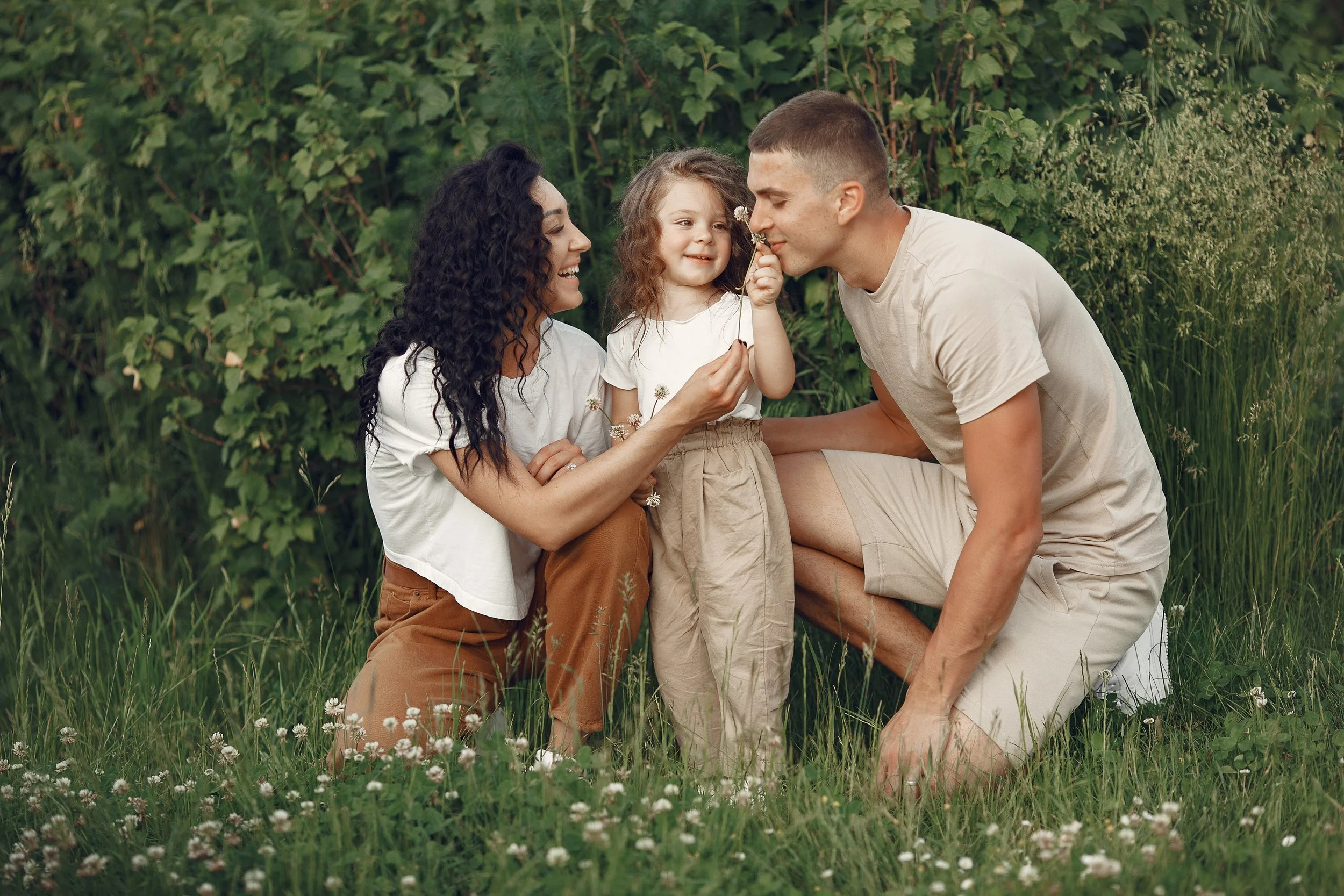 A family of three, a woman, a man, and a young girl, sitting outdoors in a grassy area with green foliage in the background. They are smiling and appear to be enjoying a moment together, with the young girl holding a small flower and the man sniffing it.
