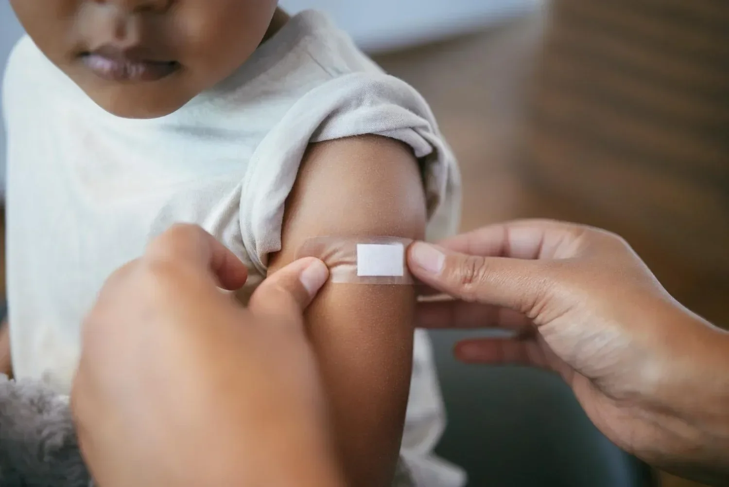 A child with a rolled-up sleeve receives a band-aid on their arm from an adult's hands, suggesting care after a vaccination.