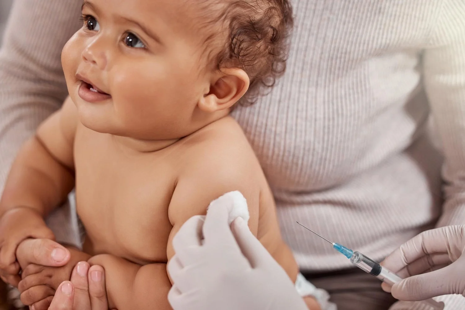 A smiling baby with curly hair receiving a vaccination injection in the upper arm from a healthcare professional wearing gloves, while sitting on an adult's lap.