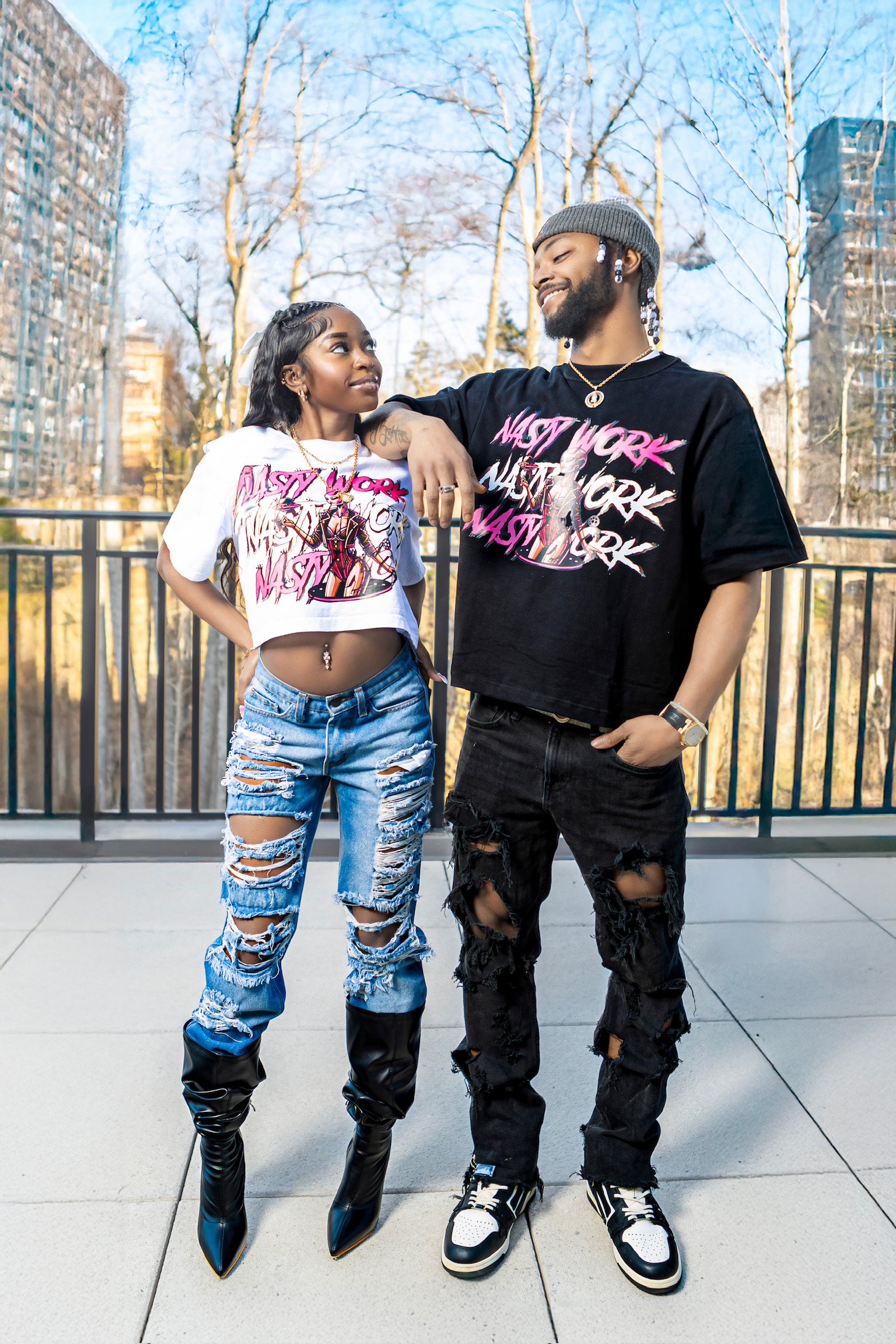 A young woman and man stand outdoors on a terrace, dressed in trendy, distressed clothing with matching 'Last Work' graphic T-shirts, looking at each other with a cityscape and trees in the background.