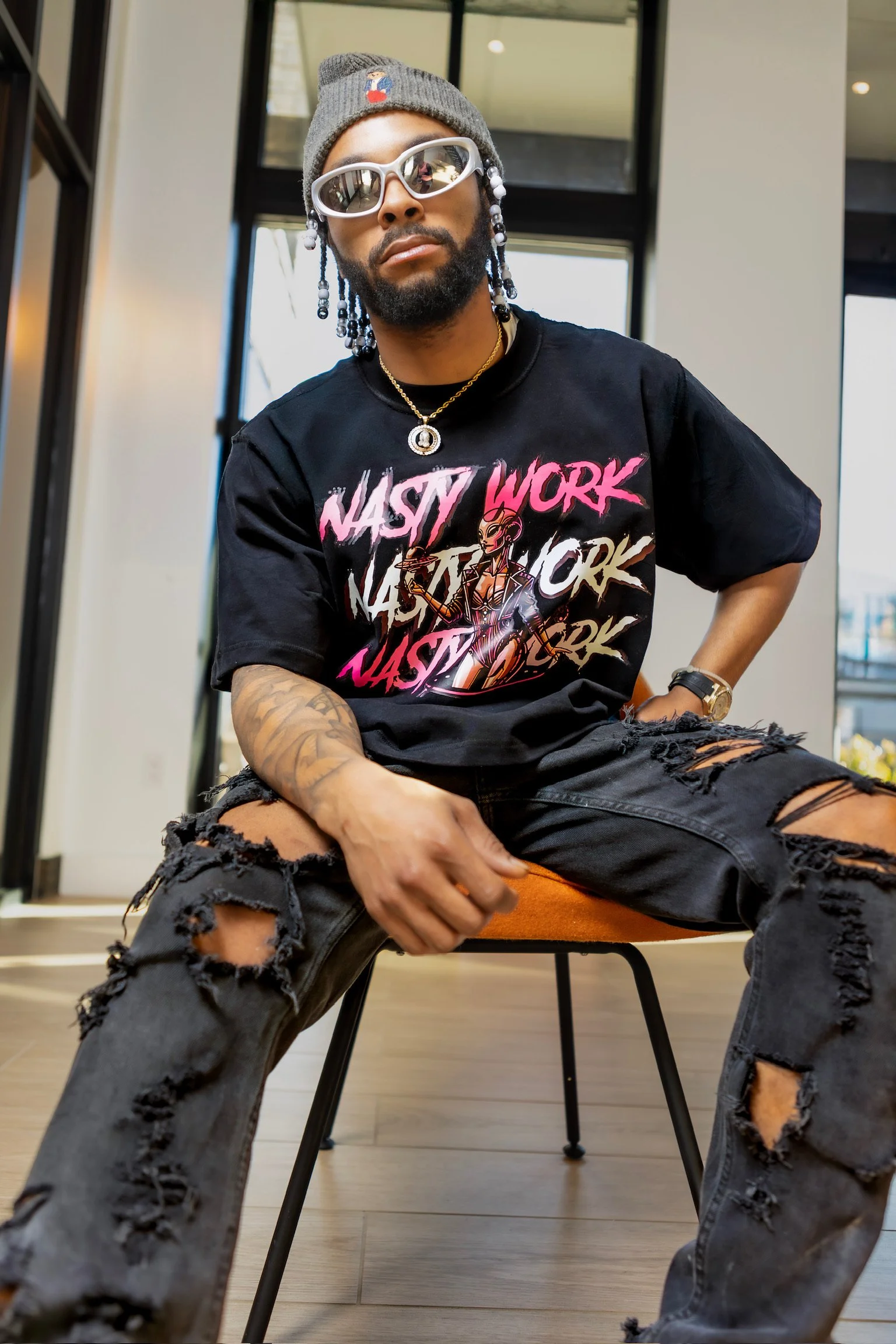 A man with a beard, sunglasses, gray beanie, and jewelry is sitting on an orange chair in a modern indoor space. He is wearing a black t-shirt with bold text and graphics and heavily distressed black jeans.