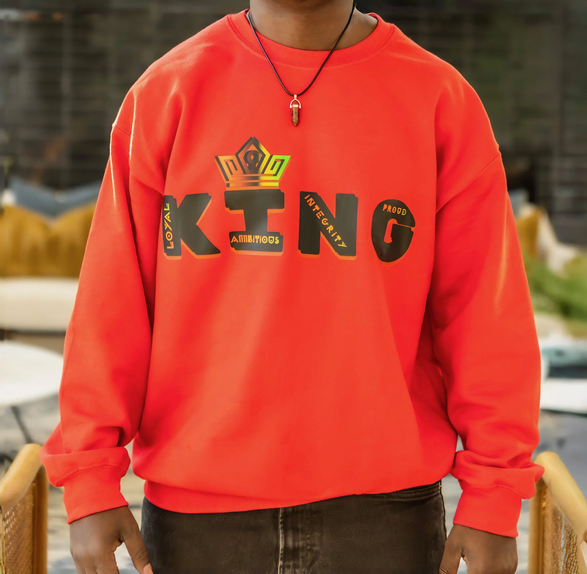 Person wearing a bright red sweatshirt with the word 'KING' in stylized lettering and words like 'Loyal', 'Ambitious', 'Integrity', and 'Proud' on the front. The person is also wearing a black necklace with a pendant.
