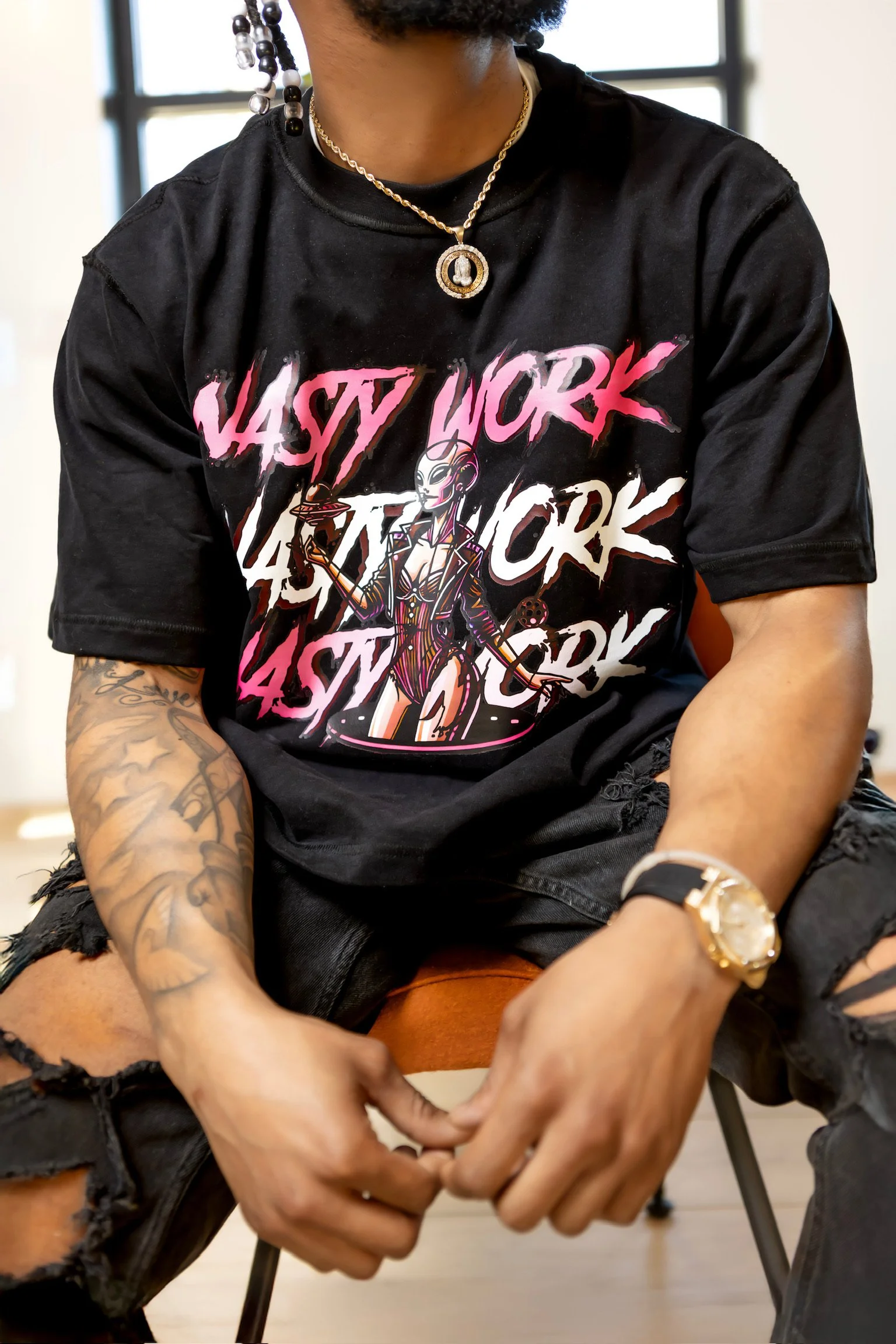 Person wearing a black graphic T-shirt with the words "NASTY WORK" repeated, paired with ripped black jeans, gold watch, layered necklaces, and earrings, sitting indoors near a window.