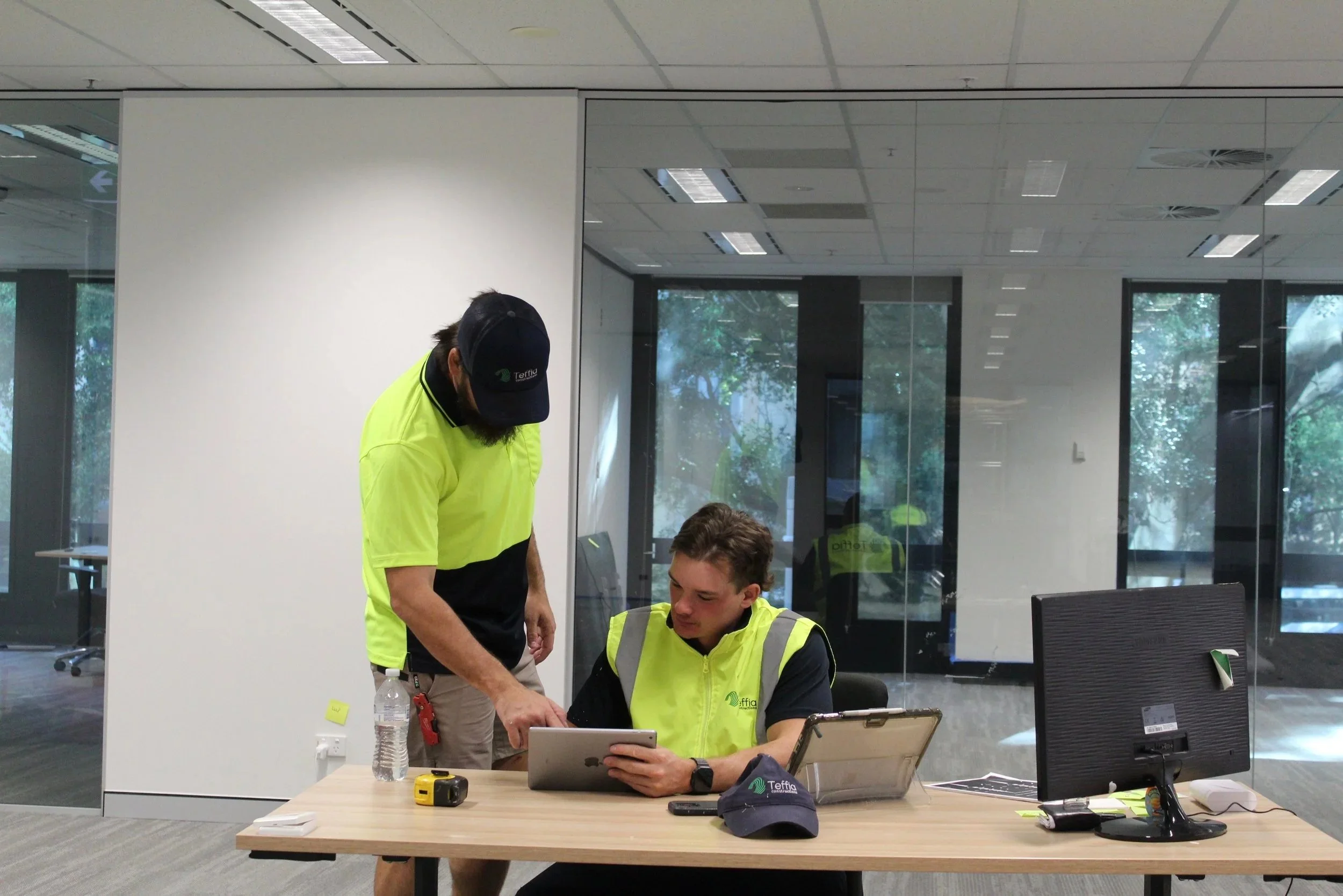 Two men in safety vests at a desk with a computer, a water bottle, and a cap, in an office with glass walls and trees outside.