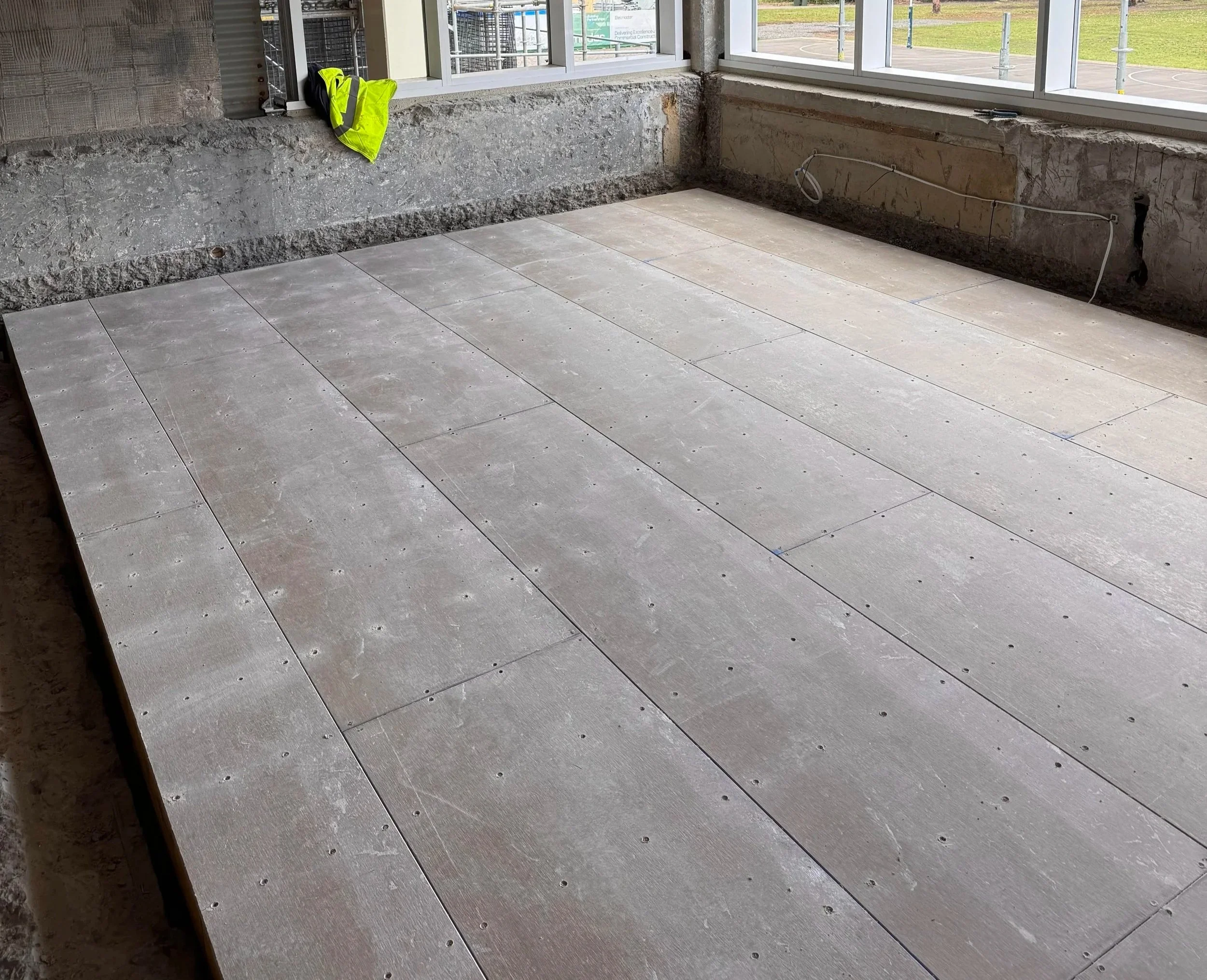 Construction site with a concrete balcony area and wooden flooring, with construction materials and tools visible. A bright yellow safety vest is hanging on a window frame.
