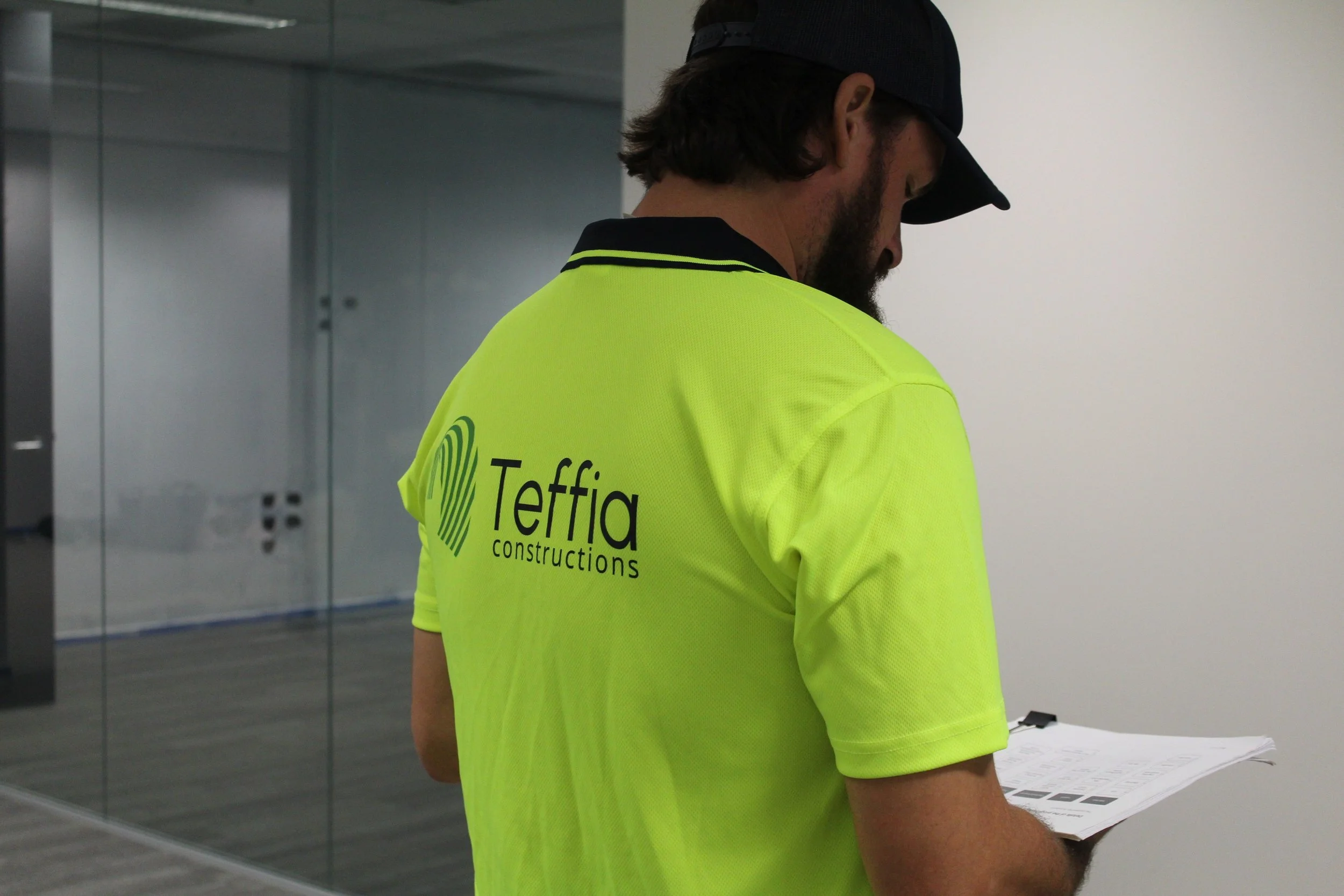 A man in a yellow safety shirt with 'Tefia Constructions' logo is holding and reading a clipboard, wearing a black cap, inside a modern office building with glass walls.