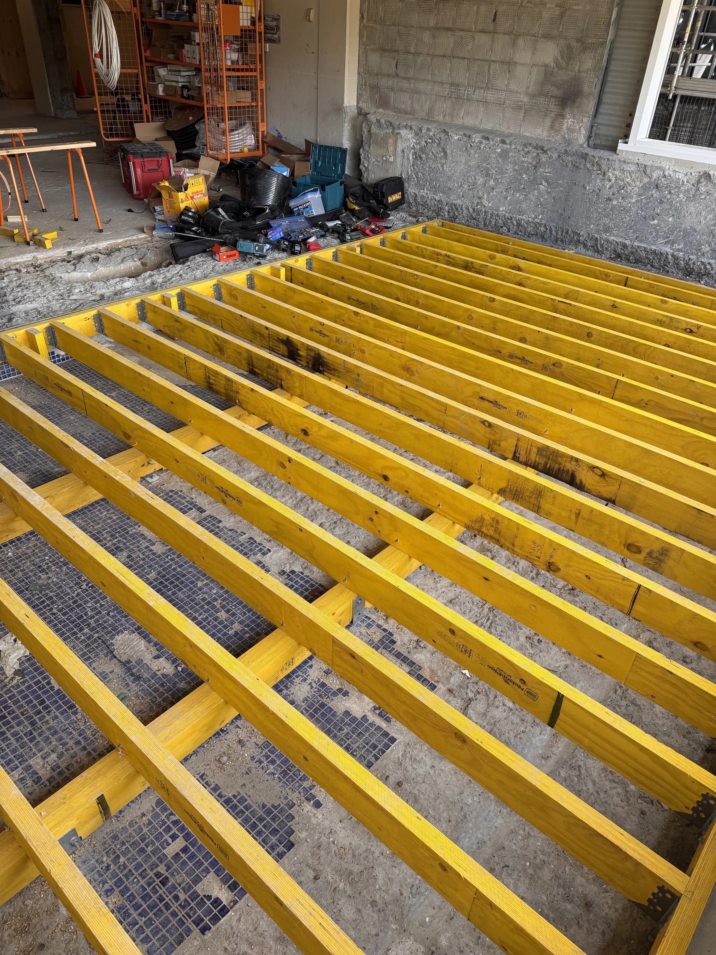 Wooden floor frame construction with yellow beams installed over a concrete subfloor, construction tools, and supplies nearby in a partially finished room.