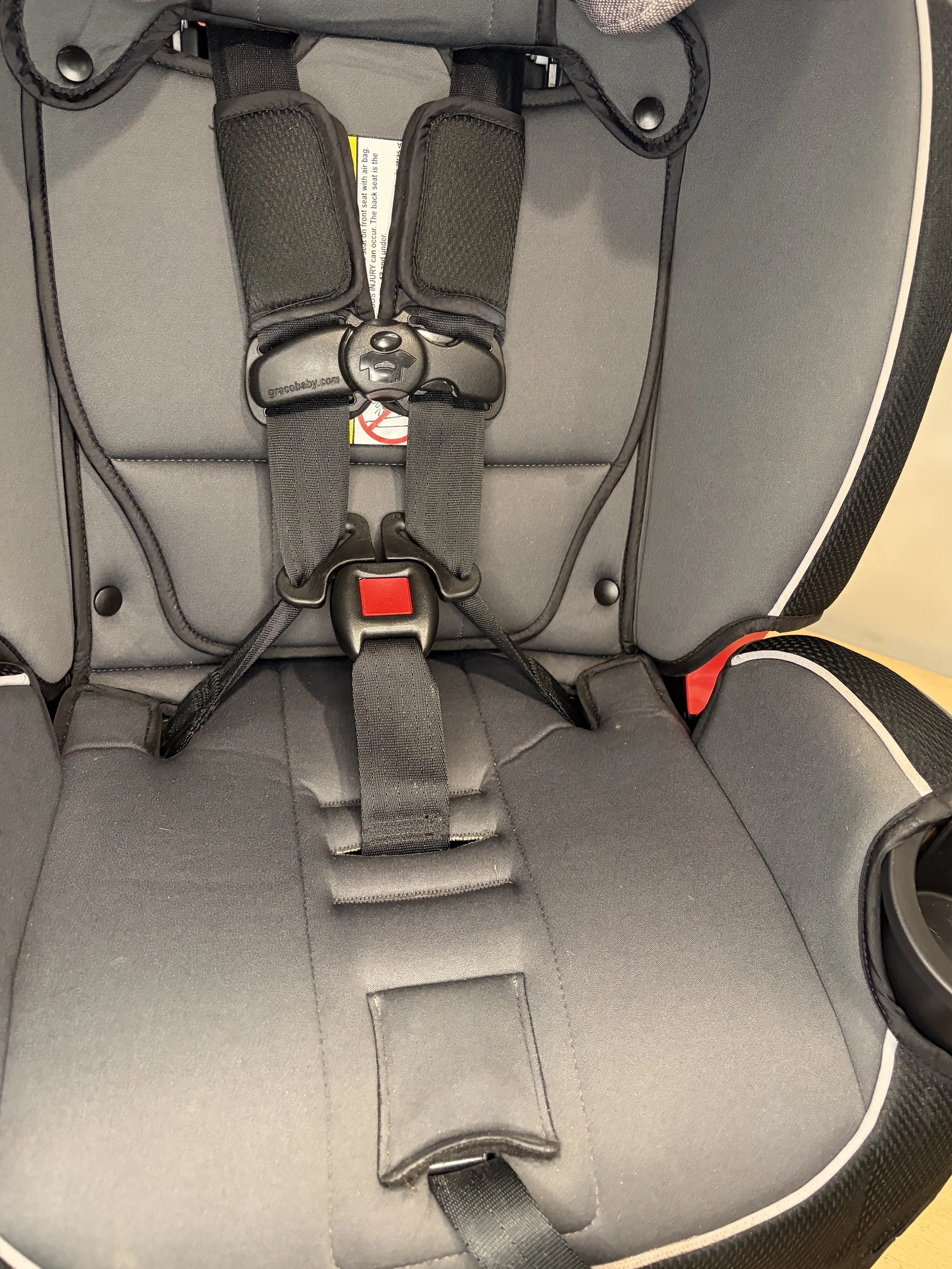 A gray child car seat with a five-point harness system, including shoulder straps, a central buckle, and a red release button, designed for safety in vehicles.