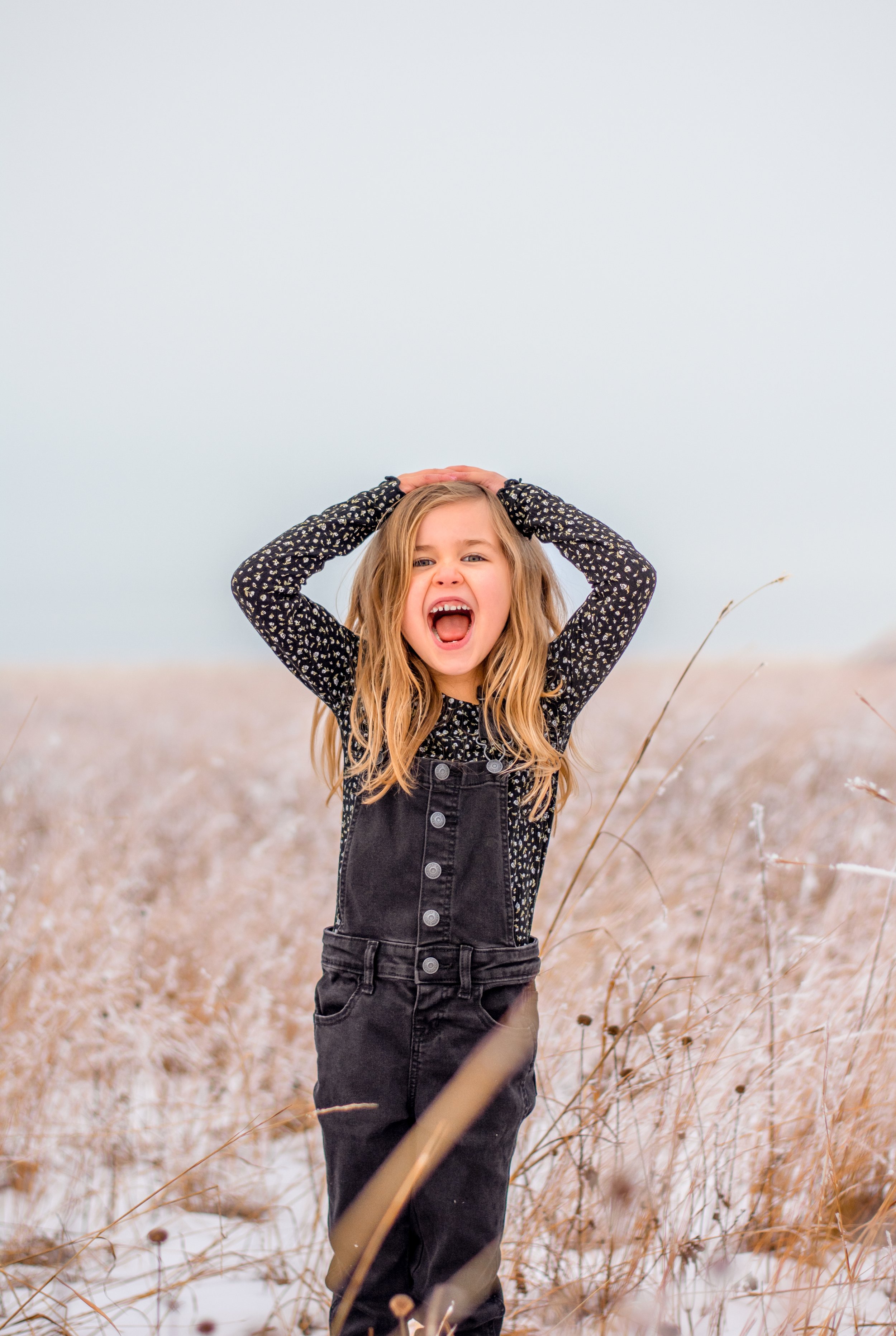 Girl with blonde hair wearing black overalls and a long-sleeve patterned shirt, standing in a snowy field with tall dry grass, yelling with her mouth open and hands on her head.