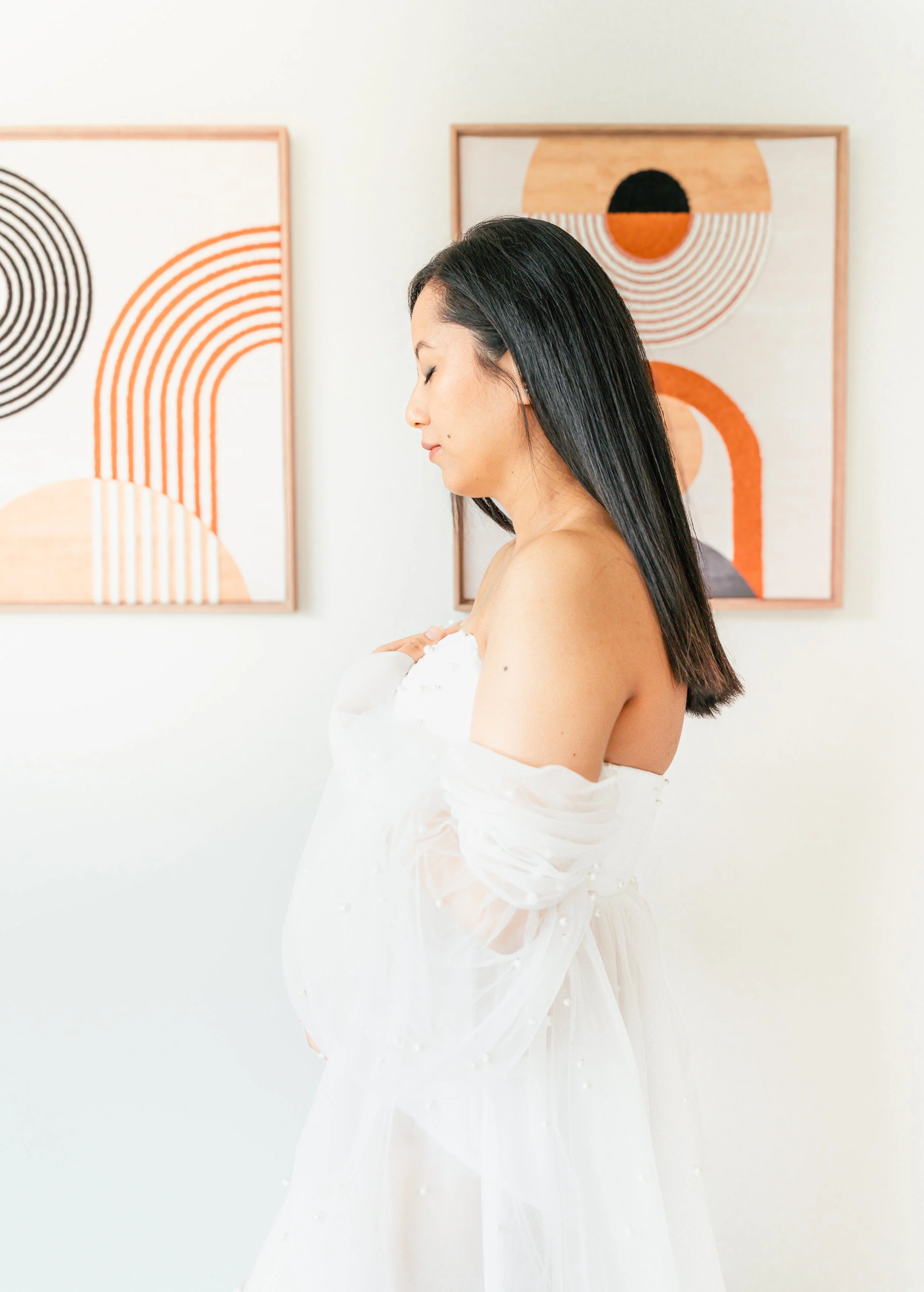 A pregnant woman with long black hair in a white dress standing in front of abstract artwork on a white wall.