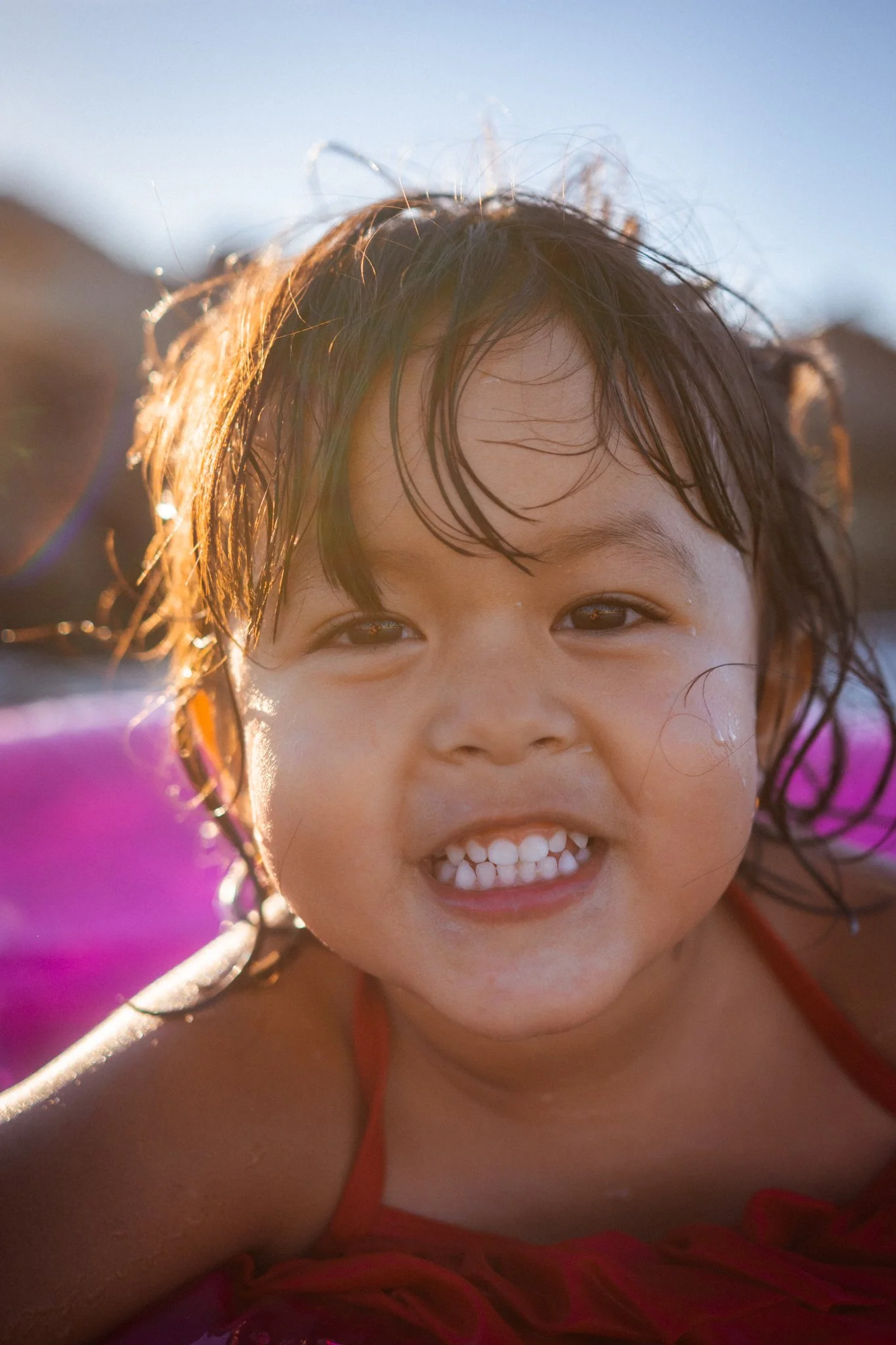 A young girl smiling with wet hair and a red swimsuit, outdoors in bright sunlight with a pink float behind her.