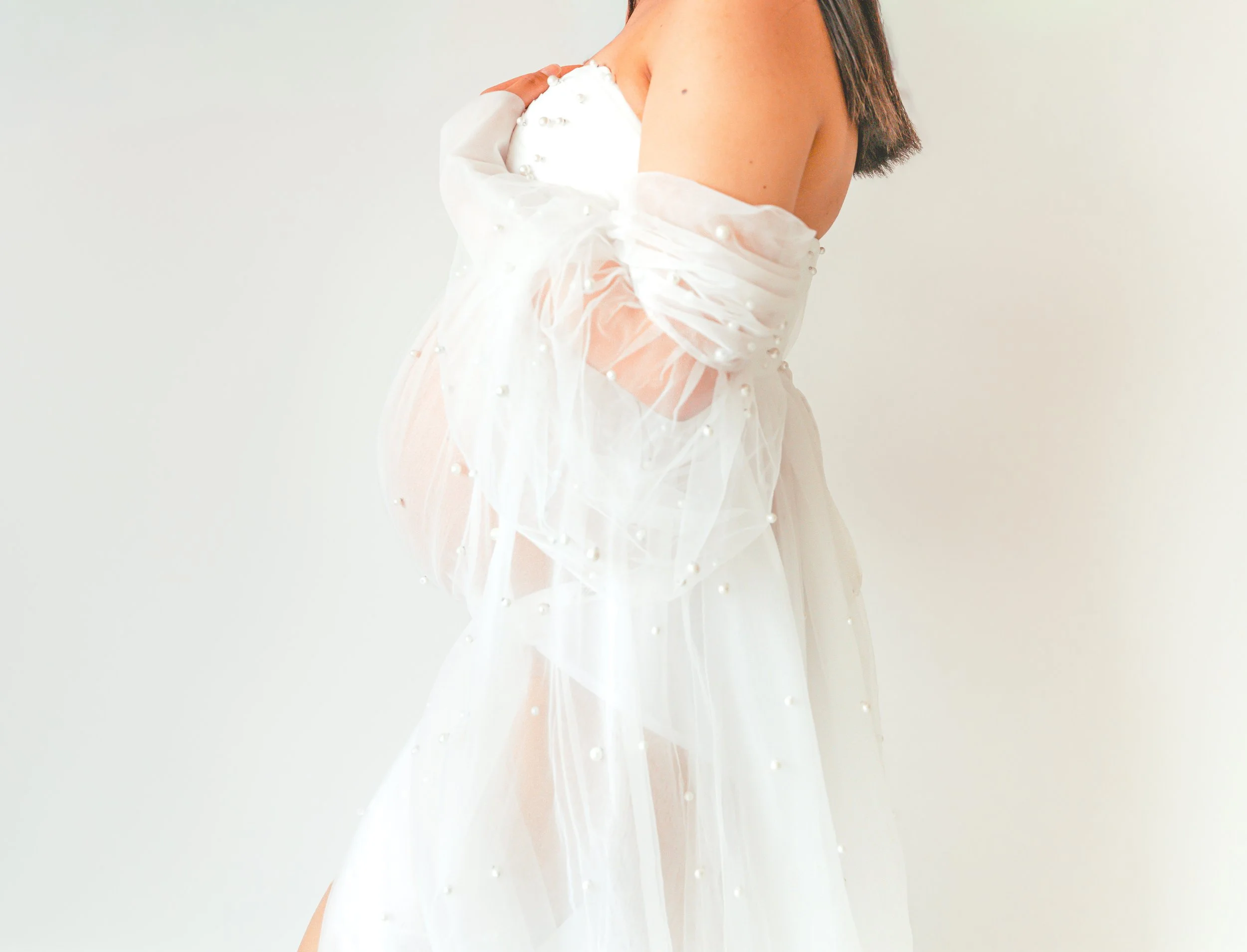 A woman in a white dress with sheer fabric and pearl embellishments, standing against a plain white background.
