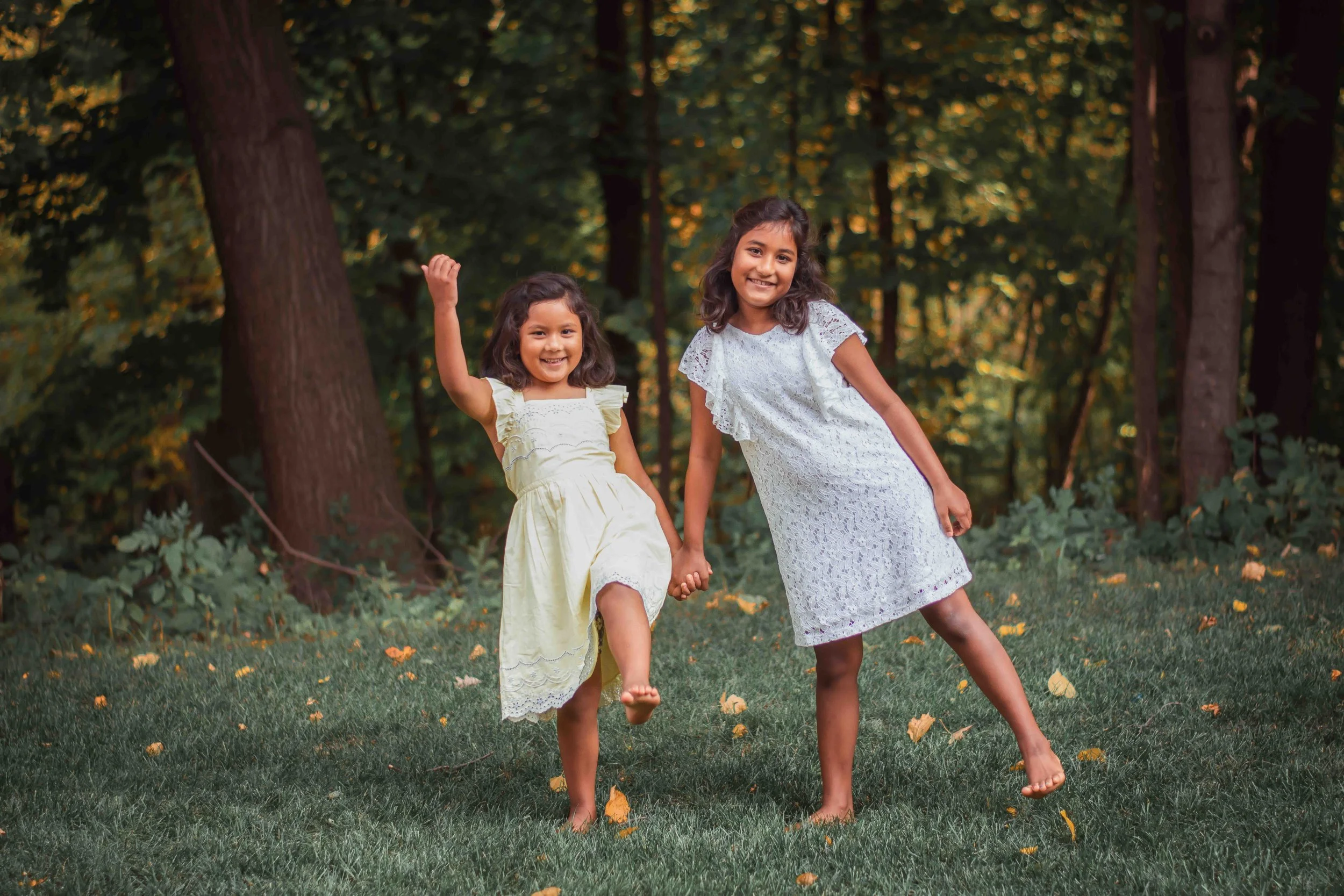 Two young girls in white dresses holding hands and playing outdoors on a grassy area surrounded by trees.