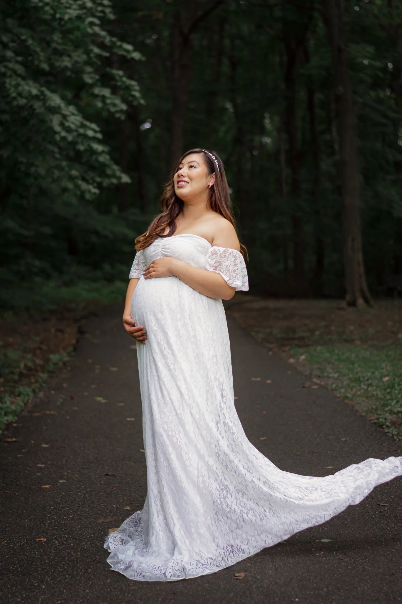 Pregnant woman in a white lace off-shoulder dress standing on a wooded path, gently cradling her belly, smiling, with trees in the background.