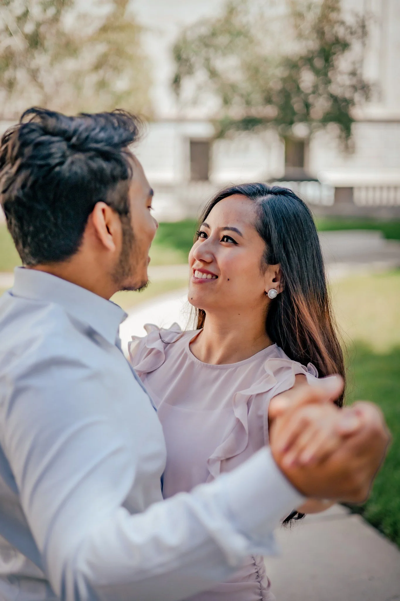 A man and woman are dancing outdoors, smiling at each other, with trees and a building in the background.