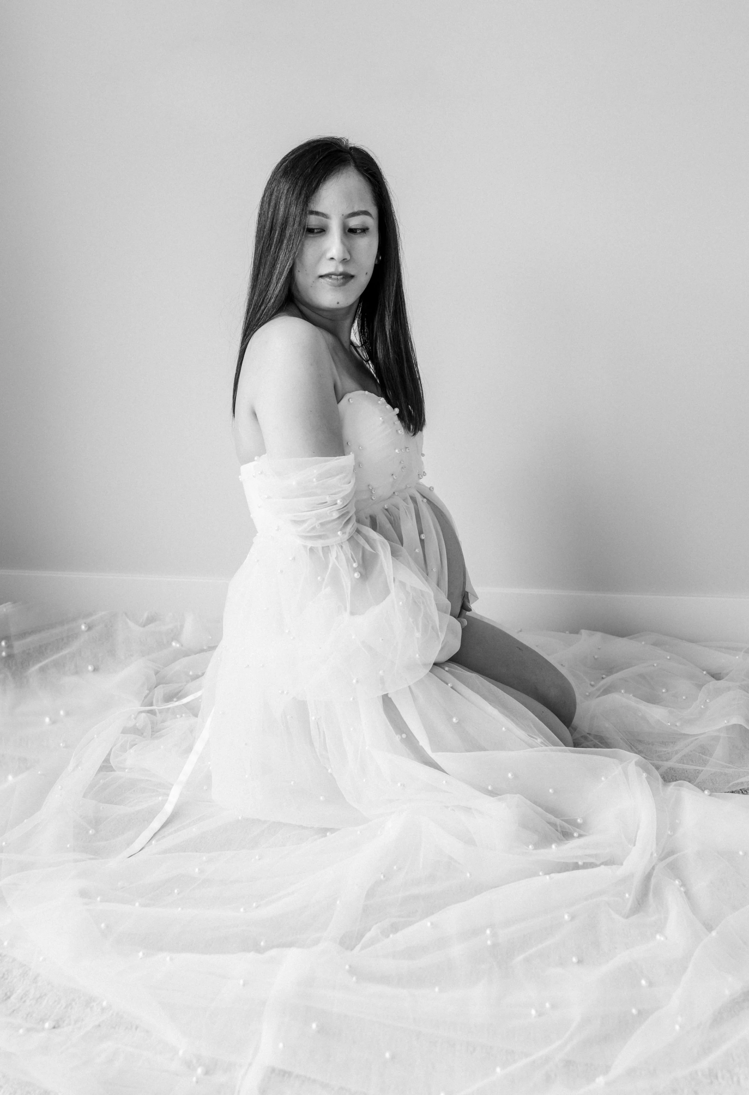 Black and white photo of a woman sitting on the floor, wearing a flowing gown with pearl embellishments, against a plain wall.