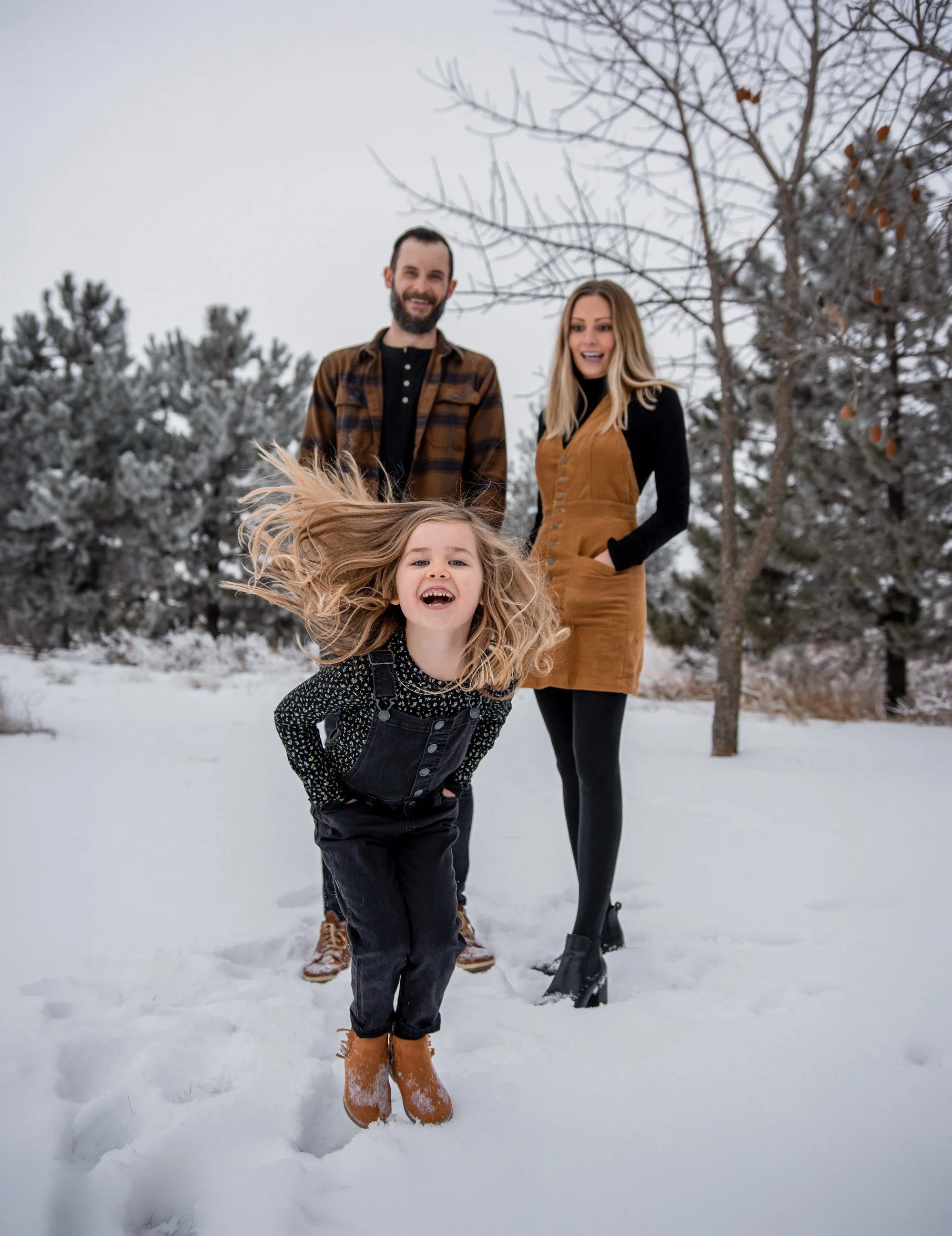 A smiling family of three, a man, a woman, and a girl, standing outside on snow-covered ground with snow-dusted trees in the background. The girl in the foreground is jumping, with loose hair and wearing brown boots, black overalls, and a black and w