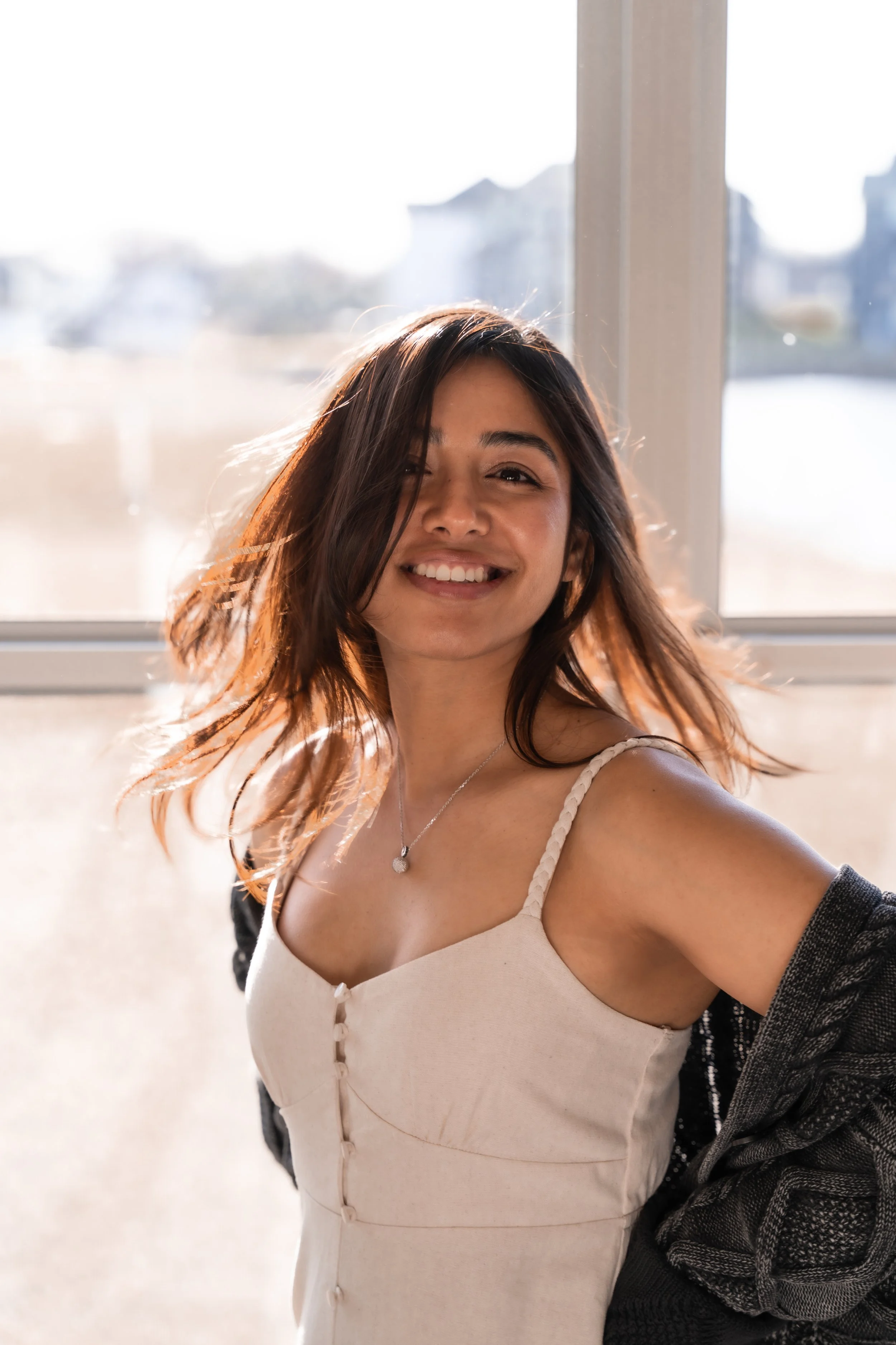 Smiling young woman with shoulder-length brown hair, wearing a white sleeveless dress and a necklace, standing in front of a window with sunlight streaming in.