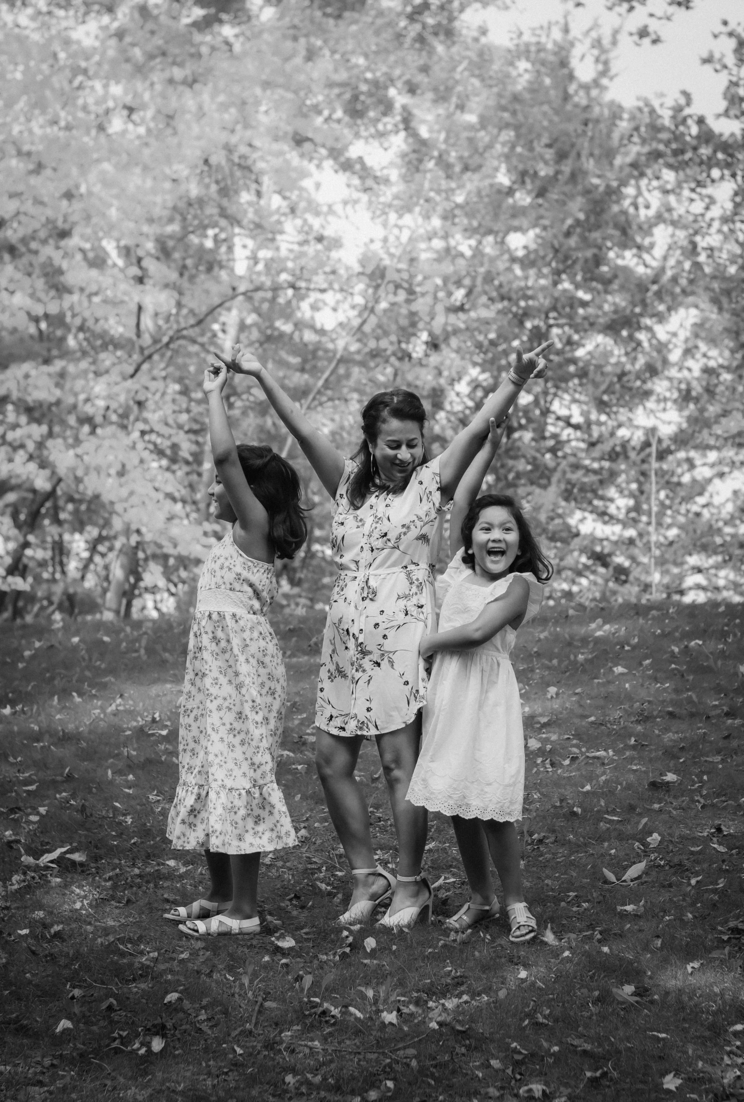 Three females, one adult and two children, joyfully dancing outdoors among trees in a park.