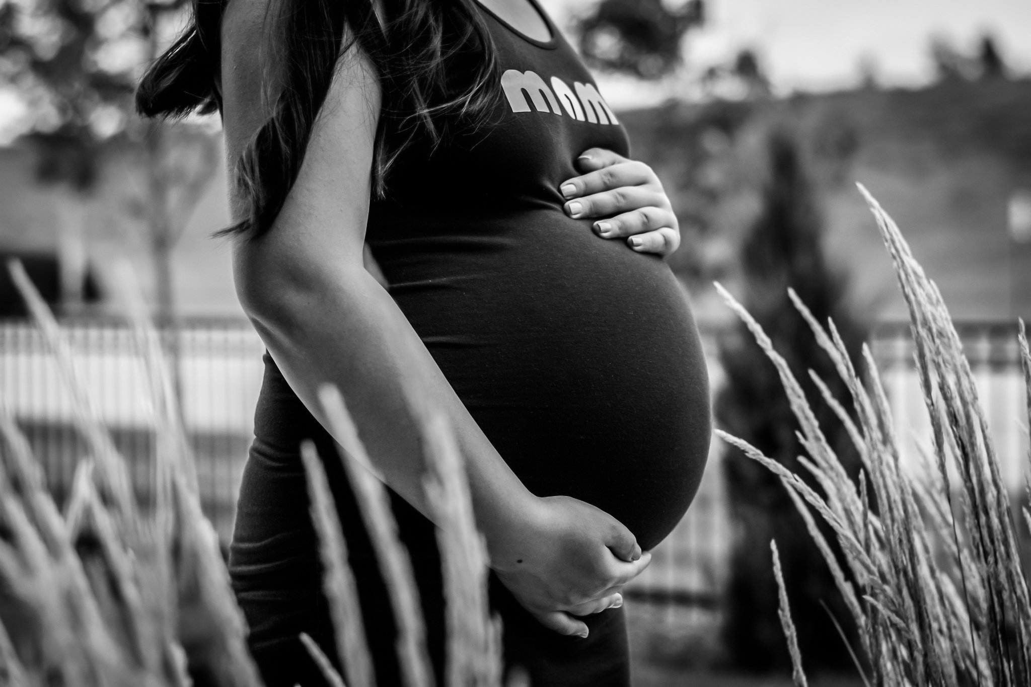 A woman with long hair wearing a black tank top and jeans is outdoors, gently cradling her pregnant belly with both hands among tall grasses, with a blurred background of trees and a fence.