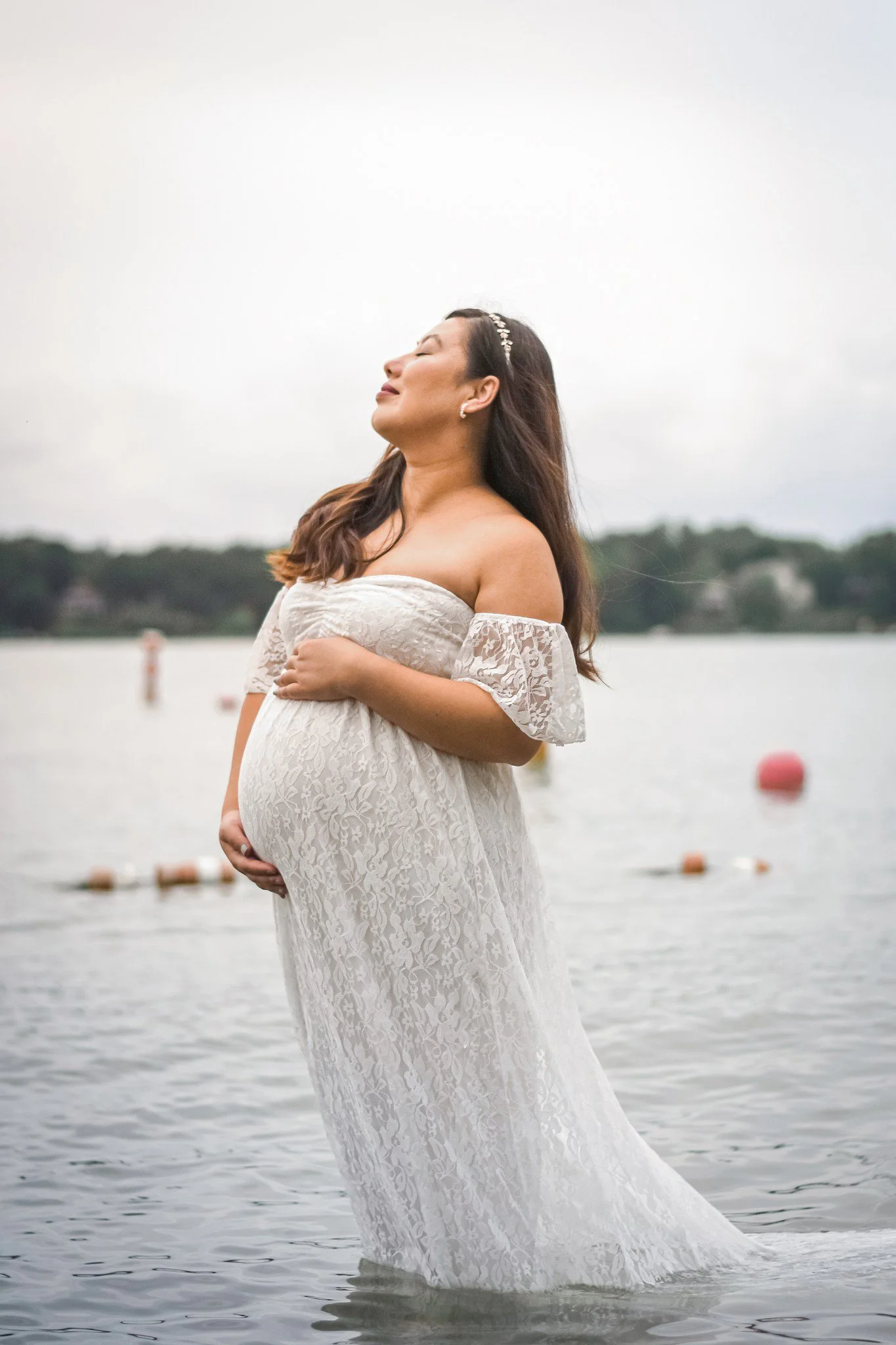 A pregnant woman in a white lace off-shoulder dress standing in shallow water by a lake, with her eyes closed and head tilted back, holding her belly with one hand.