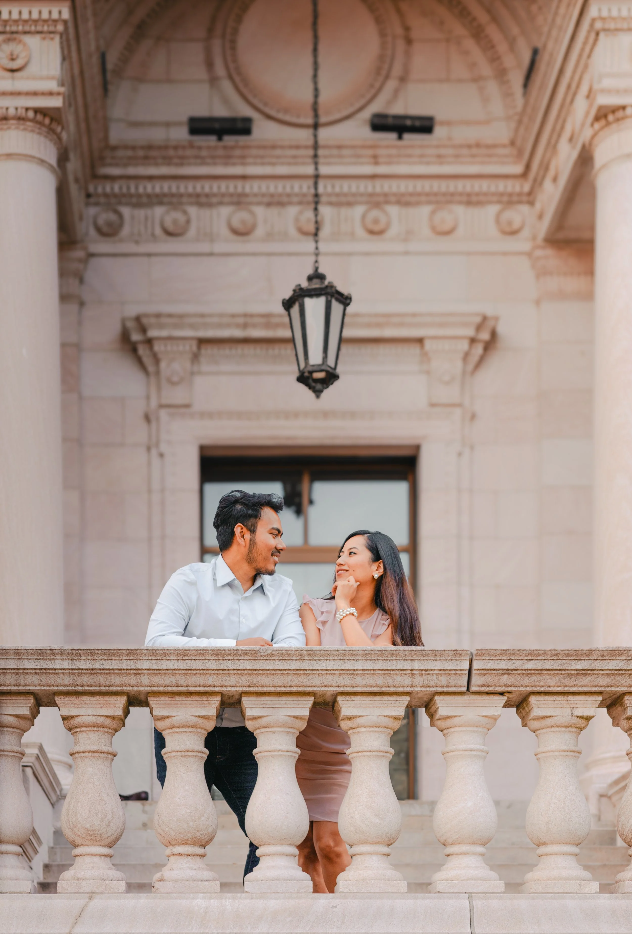A man and woman standing close on a stone balcony, smiling at each other, with a historic building and large stone columns in the background.
