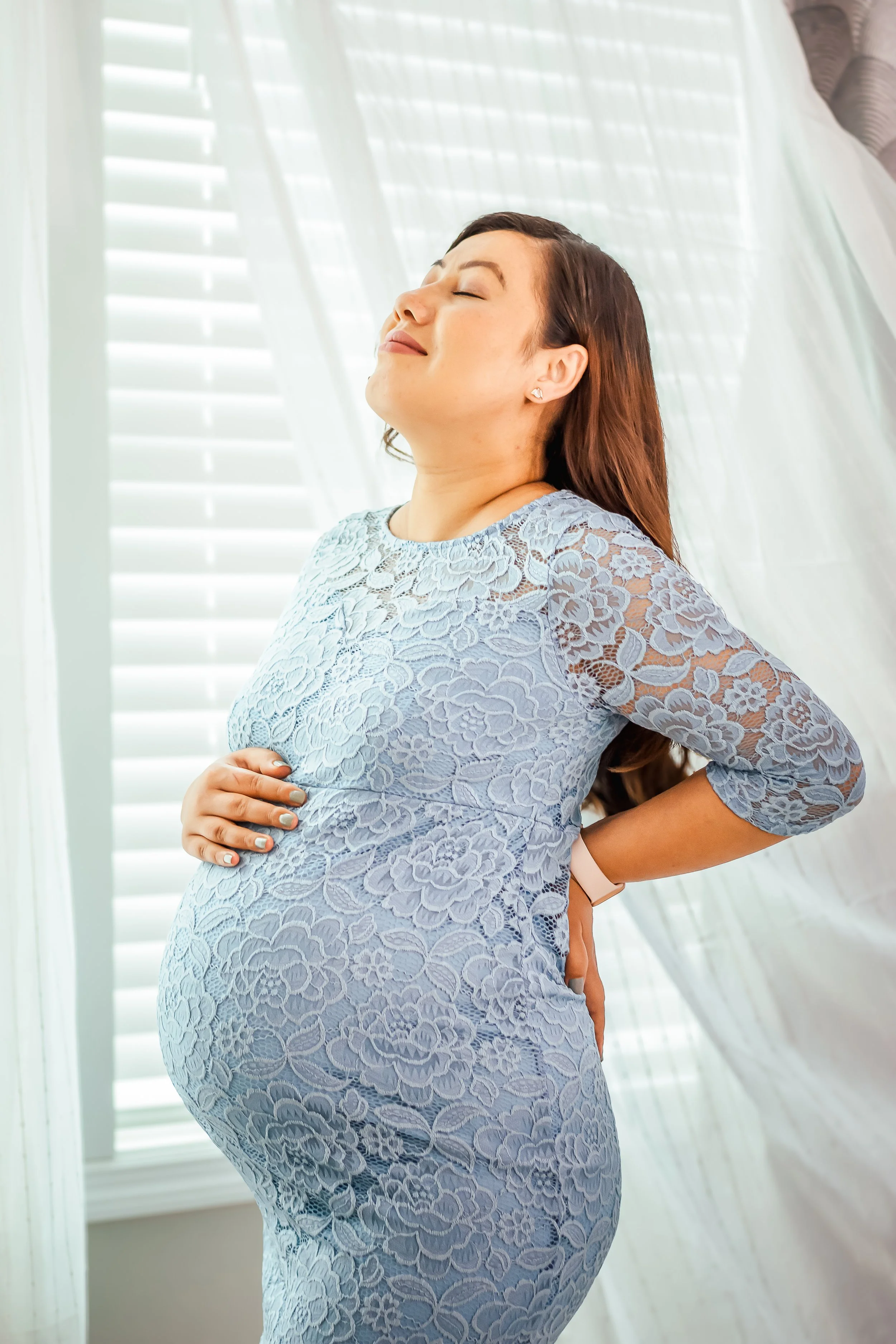 Pregnant woman in a lace dress standing with eyes closed, smiling, in front of window blinds with sunlight.