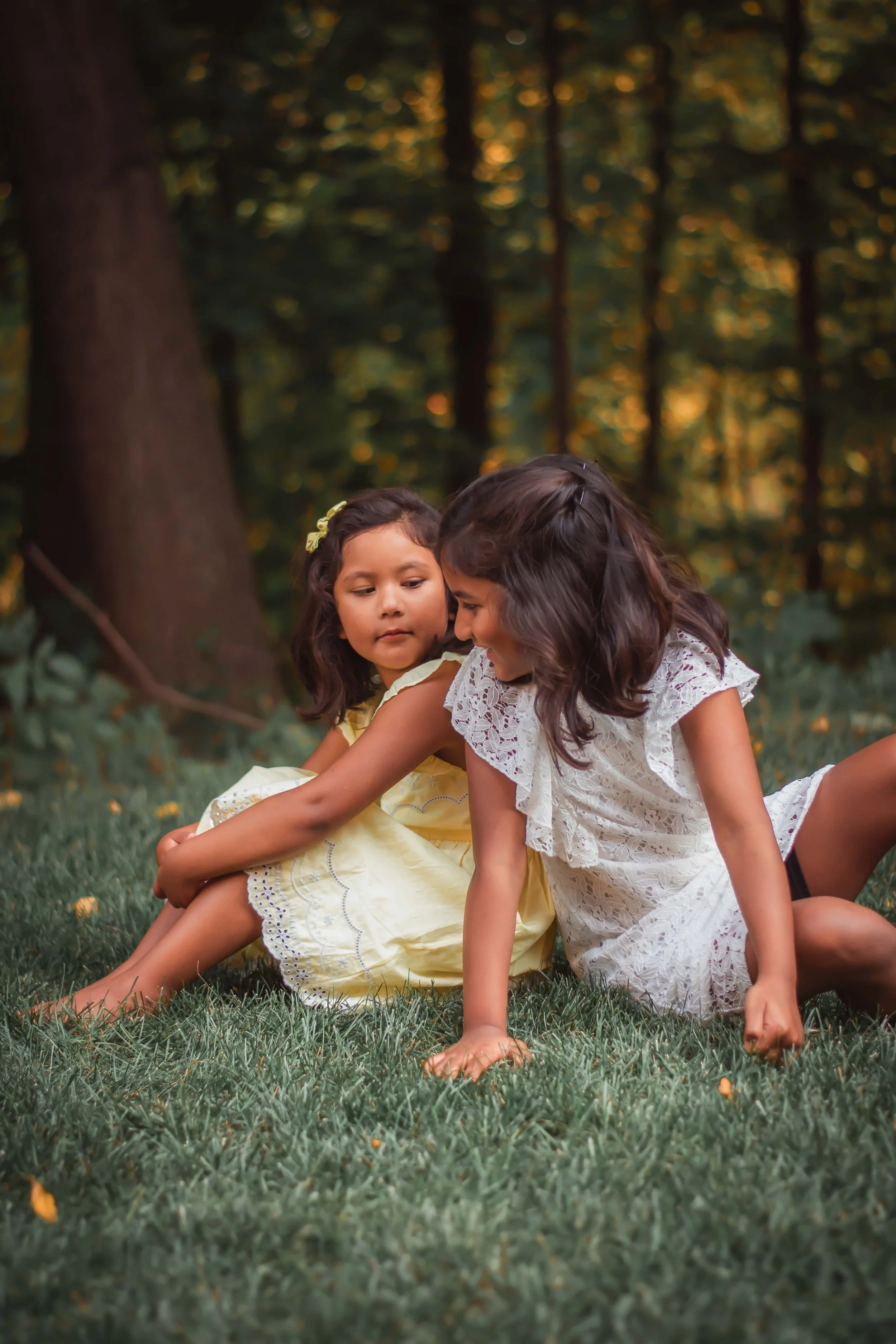Two young girls in white and yellow dresses sitting on grass in a wooded area.
