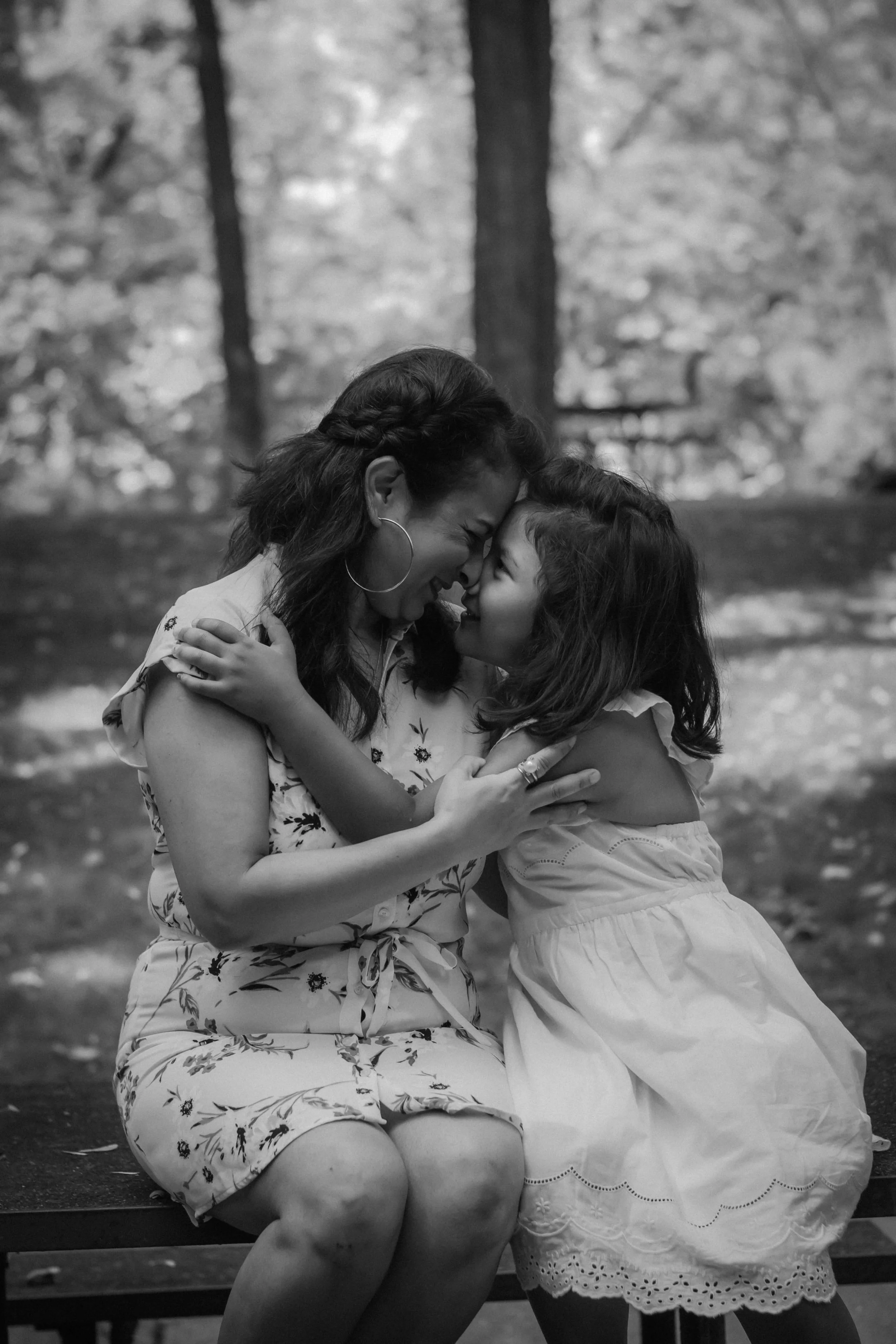 A woman and a girl sharing an embrace and touching foreheads on a park bench, smiling, black and white photo.