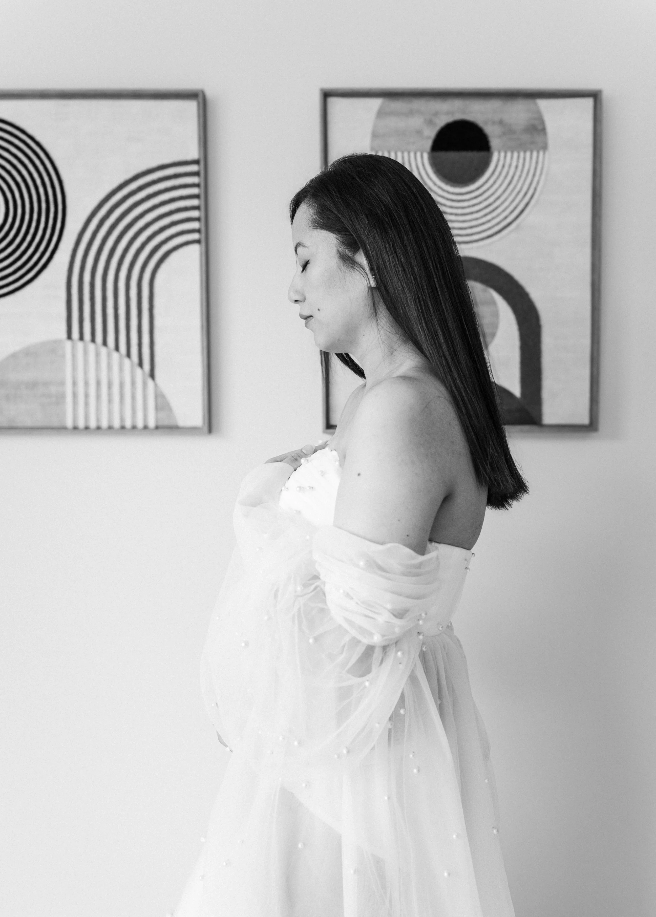 A side profile of a woman with long dark hair, wearing a white strapless dress with tulle and pearl embellishments, standing indoors in front of abstract geometric artwork.