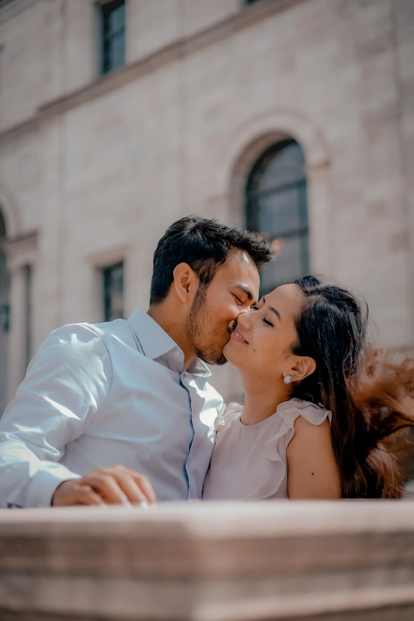 A young couple sharing an intimate kiss outdoors in front of a historic building with arched windows.