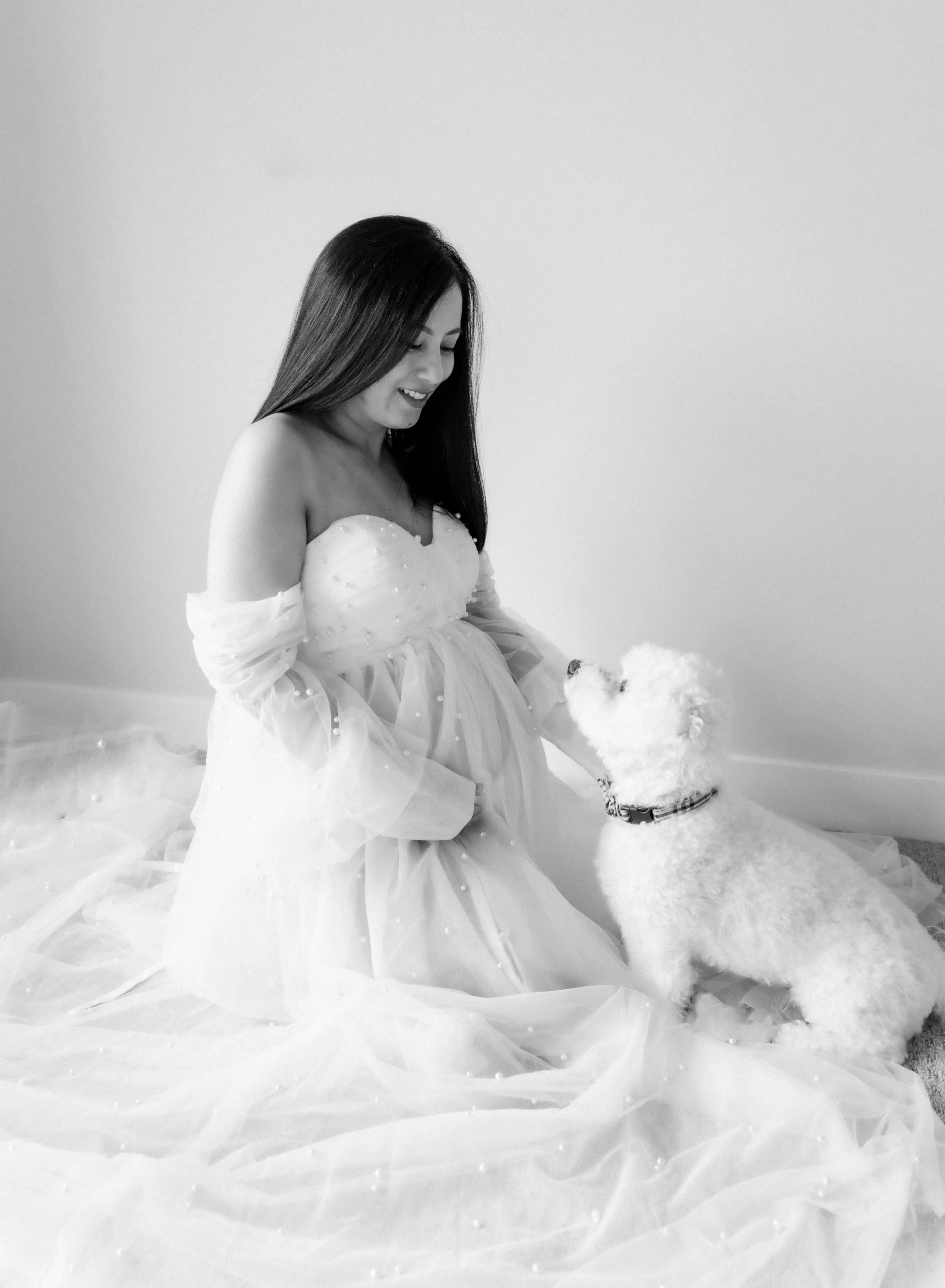 A woman in a wedding dress sitting on the floor and smiling at a small white poodle dog.